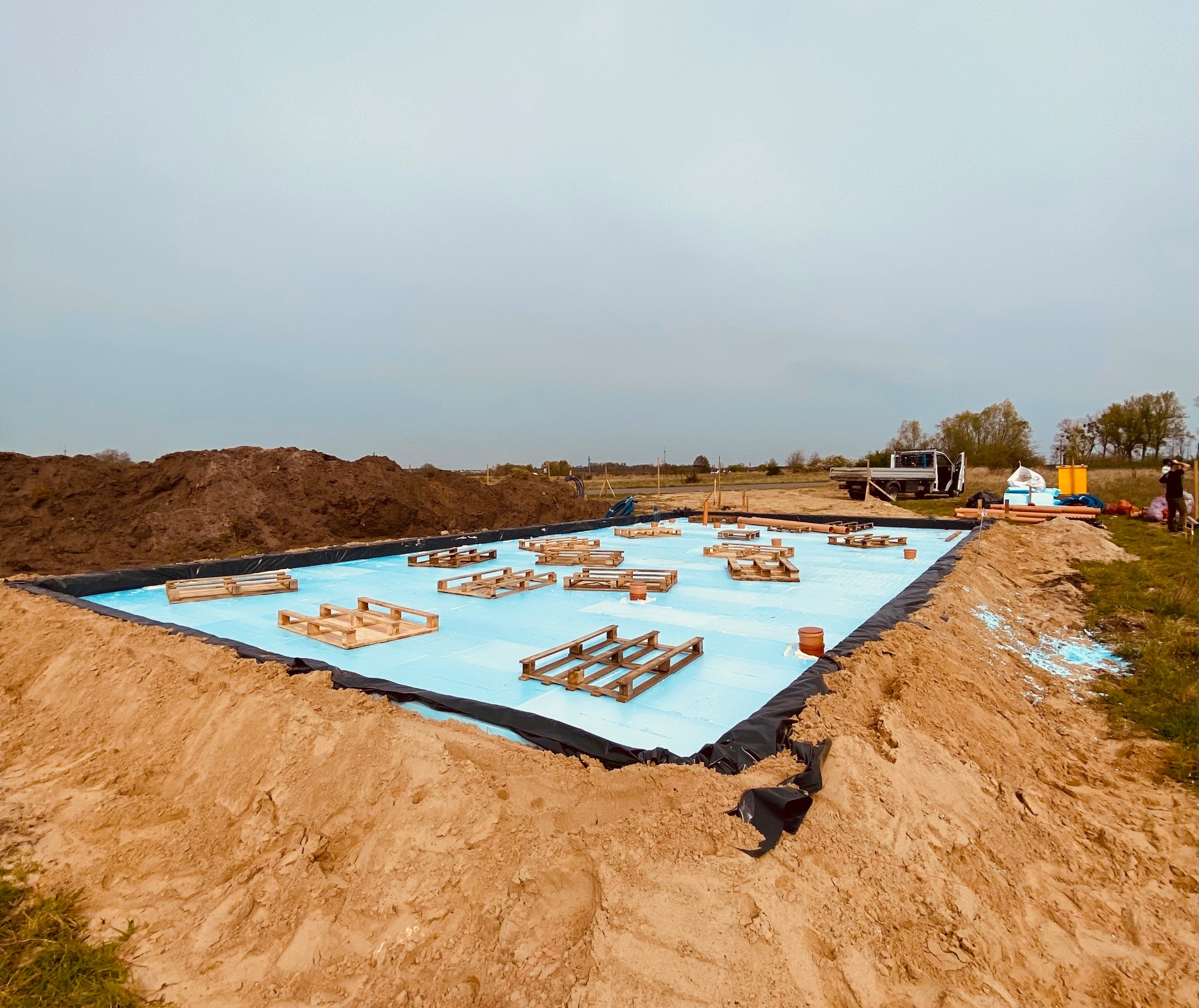 Przygotowanie podłoża pod fundamenty: izolacja z niebieskiego styropianu zabezpieczona czarną folią, drewniane palety rozłożone na powierzchni, otoczone wykopaną ziemią, widoczny samochód dostawczy...