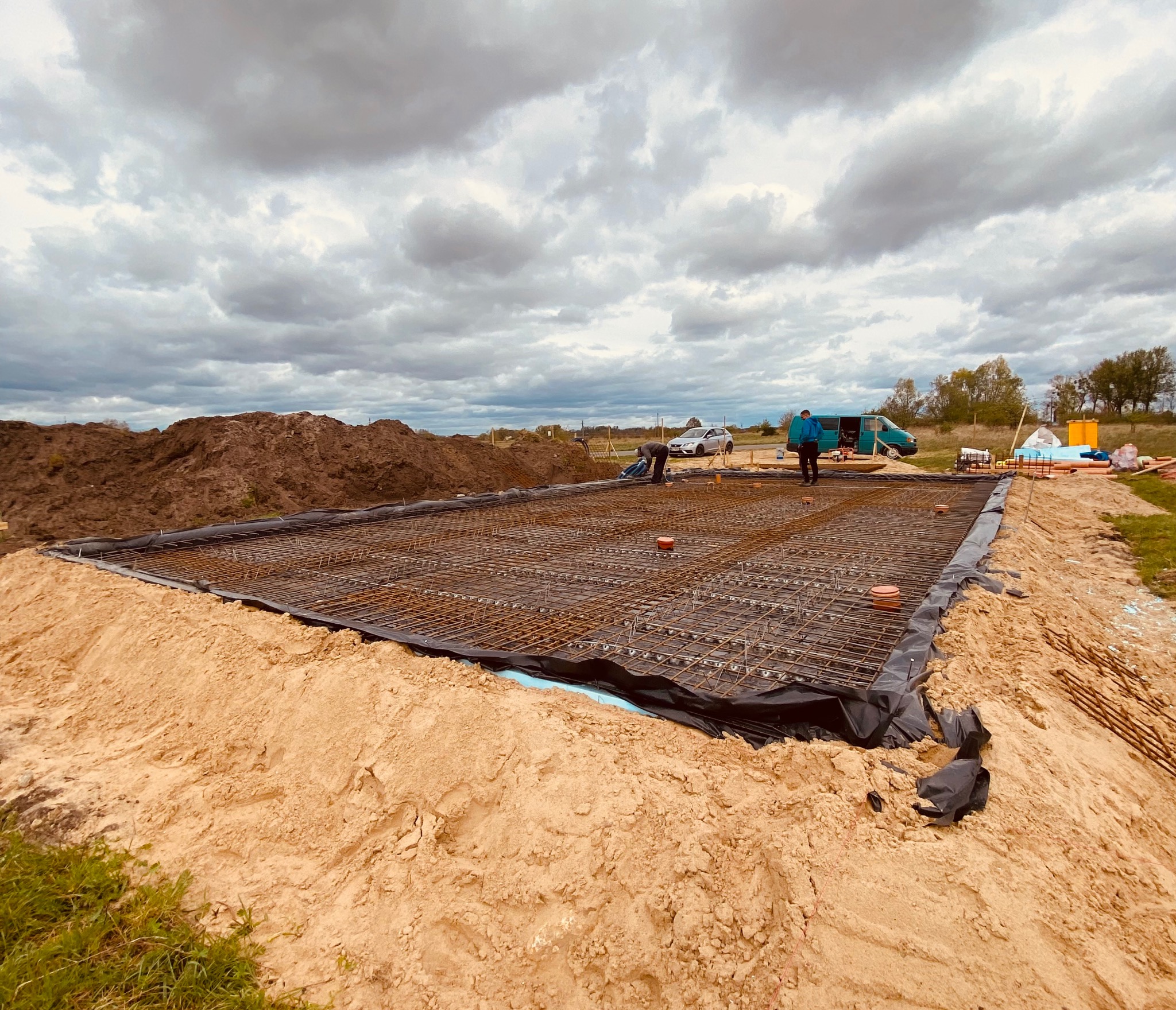 Zbrojenie płyty fundamentowej na czarnej folii, widoczne wystające pręty, pomarańczowe elementy dystansowe, dwóch pracowników w tle, hałda ziemi i pojazdy na działce budowlanej pod pochmurnym niebem.