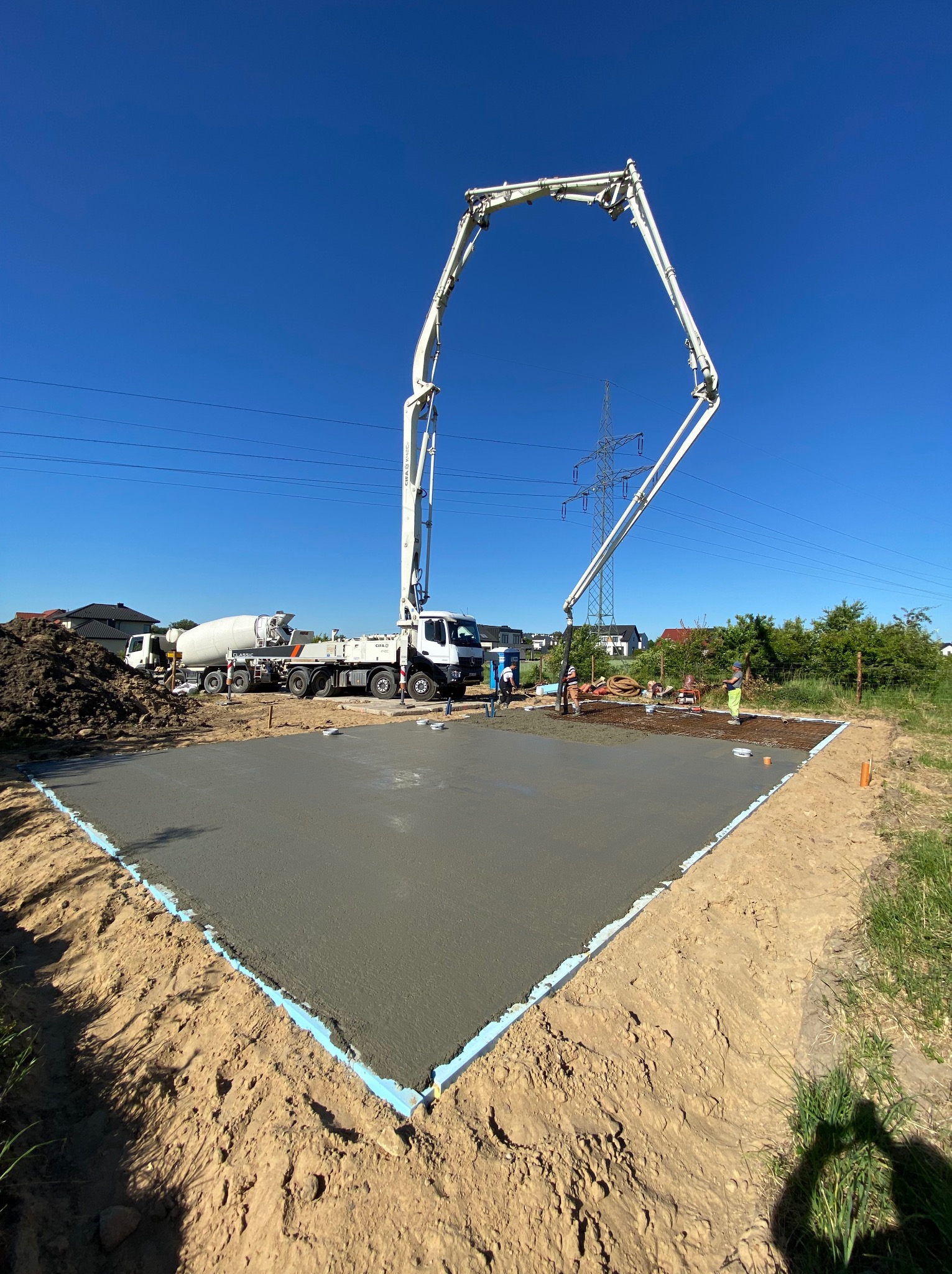 Wylewanie betonu na przygotowany teren pod fundamenty z użyciem pompy do betonu; widoczny mikser betonu, pracownicy w odblaskowych ubraniach i niebieska izolacja obwodowa.