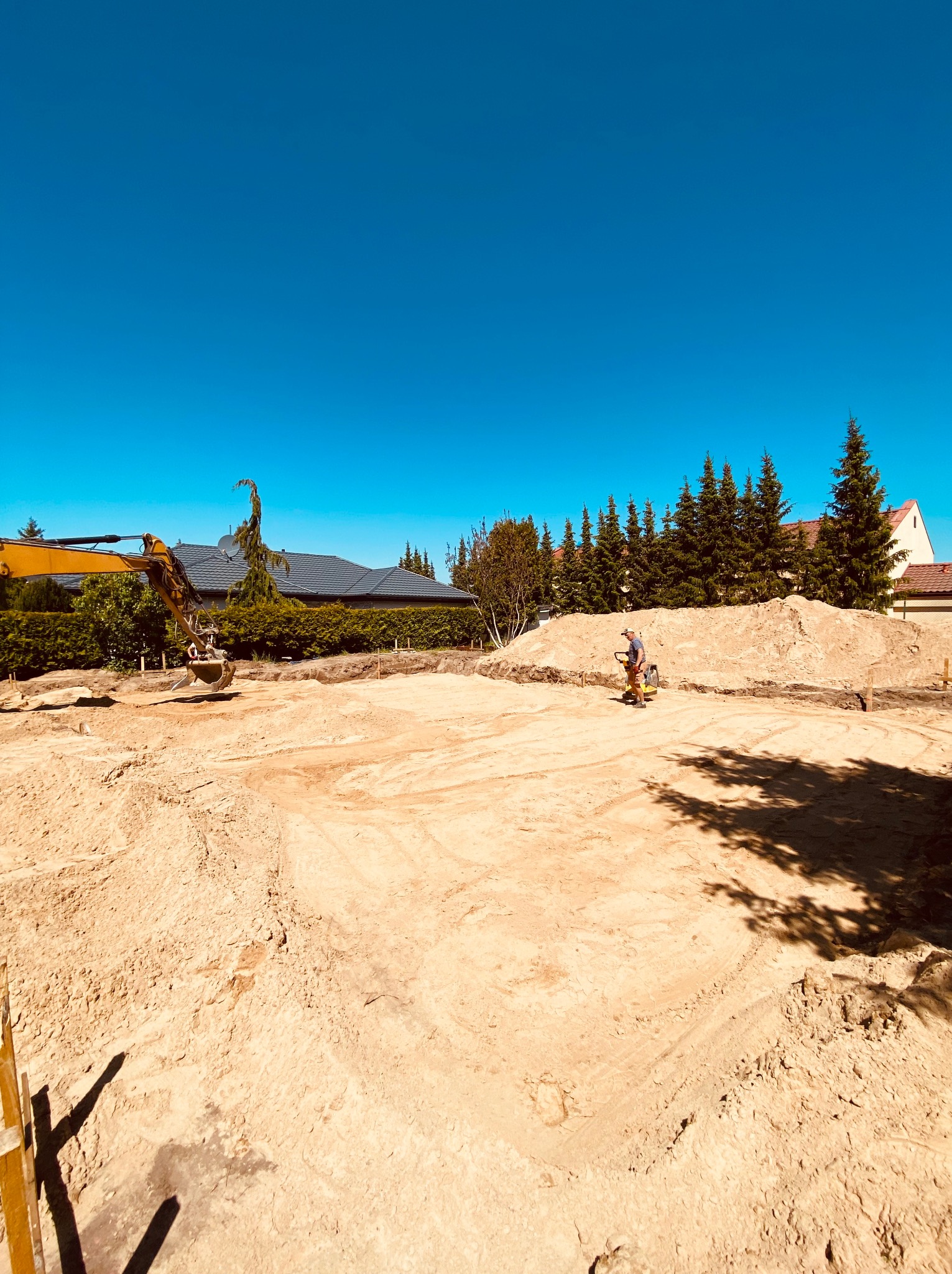 Żółta koparka i ubijarka w akcji na placu budowy, przygotowany teren pod budowę domu z widocznymi pryzmami piasku i drzewami w tle.