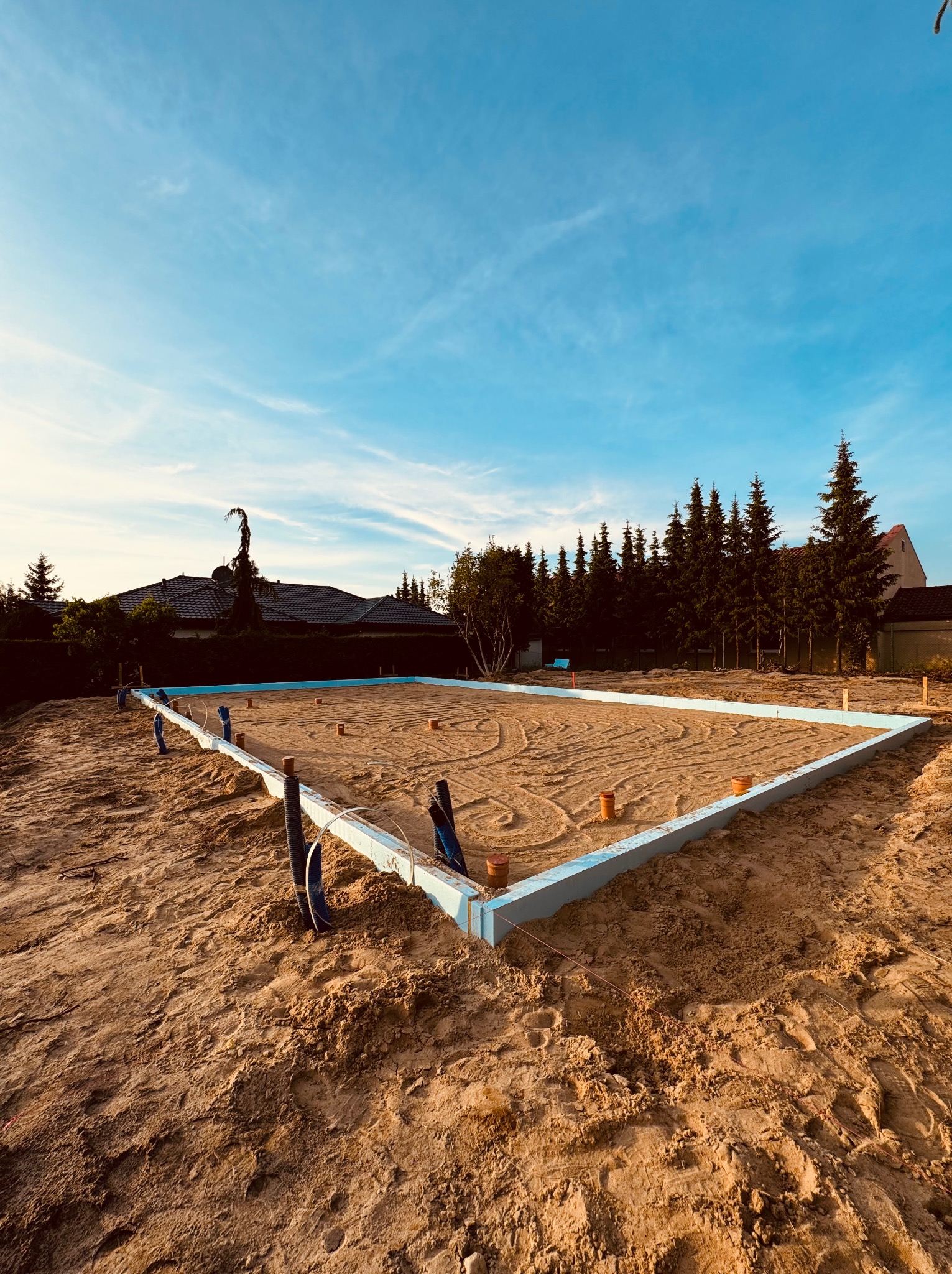 Przygotowany teren pod budowę domu z oznaczonym obrysem fundamentów, widoczne rury instalacyjne i wyrównany piasek, w tle zabudowa jednorodzinna i drzewa.