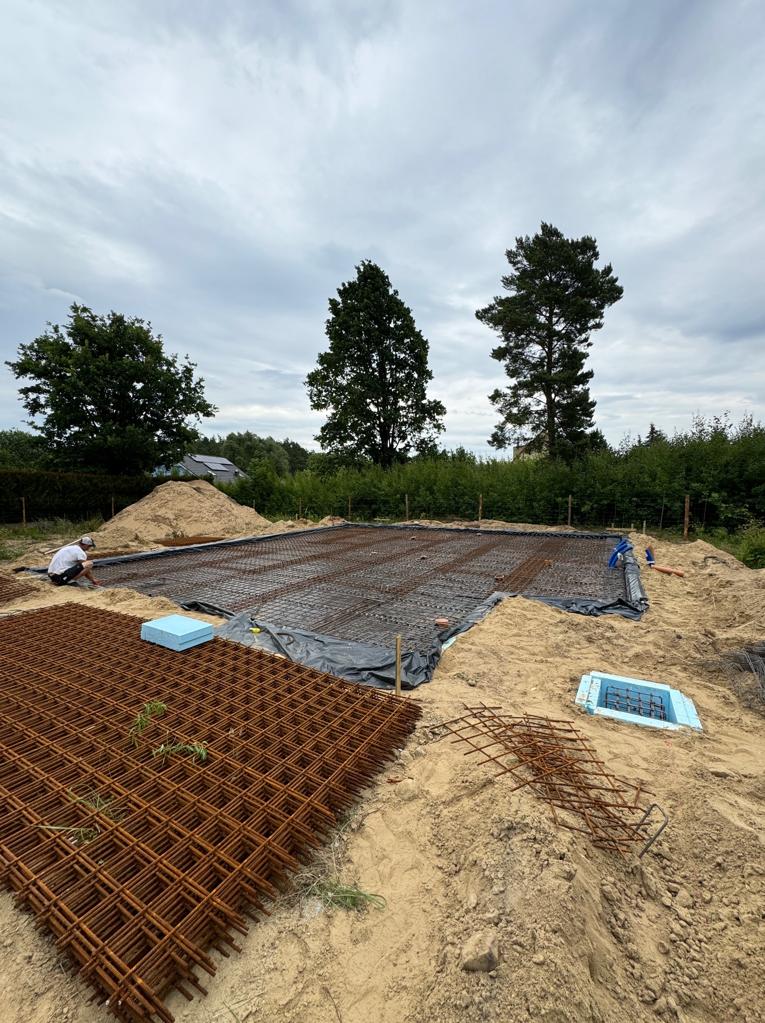 Przygotowanie zbrojenia płyty fundamentowej na gruncie pokrytym czarną folią, widoczny pracownik w trakcie montażu, ułożone siatki zbrojeniowe, niebieski styropianowy szalunek i narzędzia...