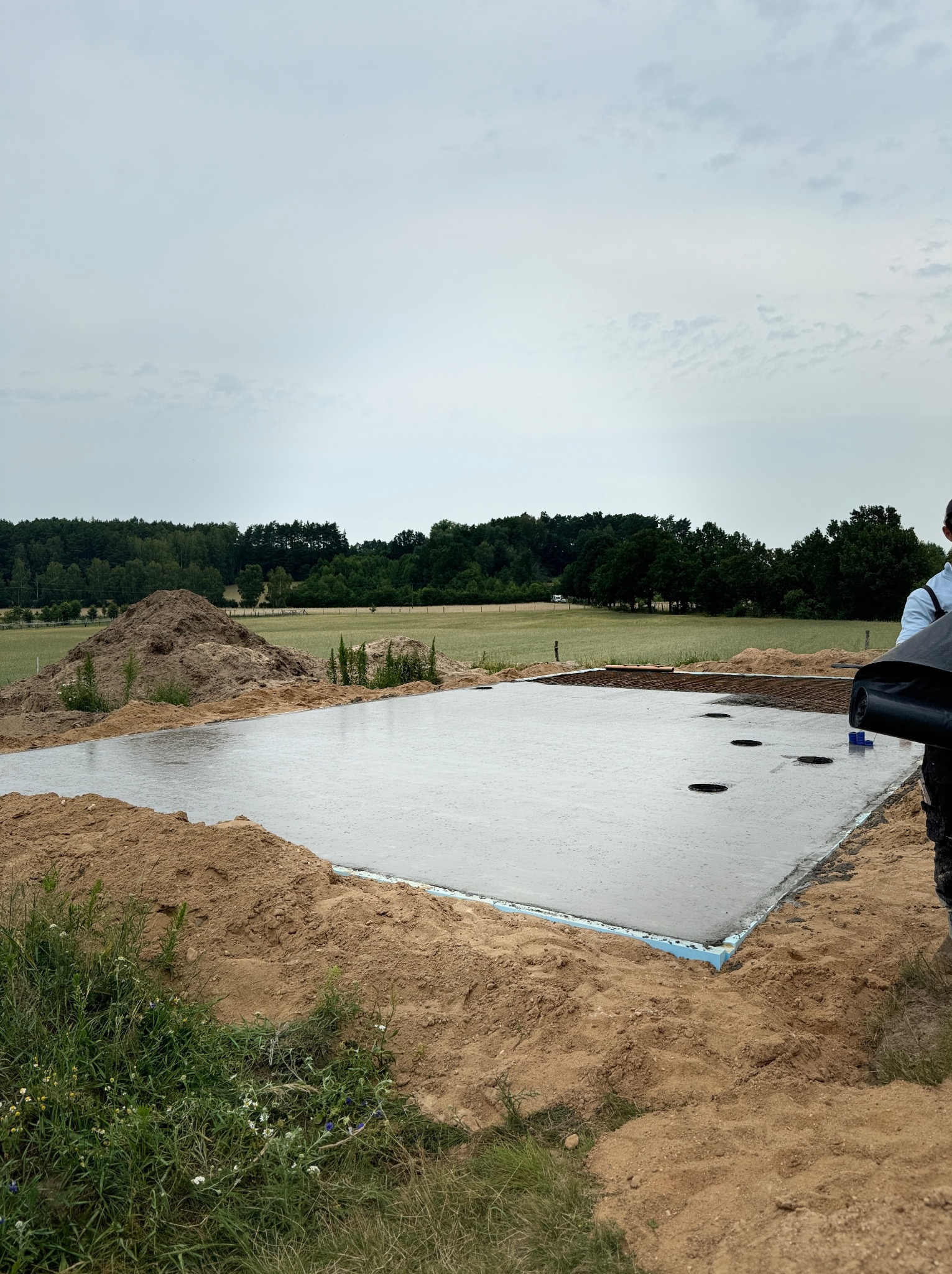 Wylany beton na przygotowanym podłożu pod fundamenty domu jednorodzinnego, widoczne izolacje termiczne, stalowe zbrojenia i wycięte otwory na instalacje, w tle zielona łąka i drzewa.