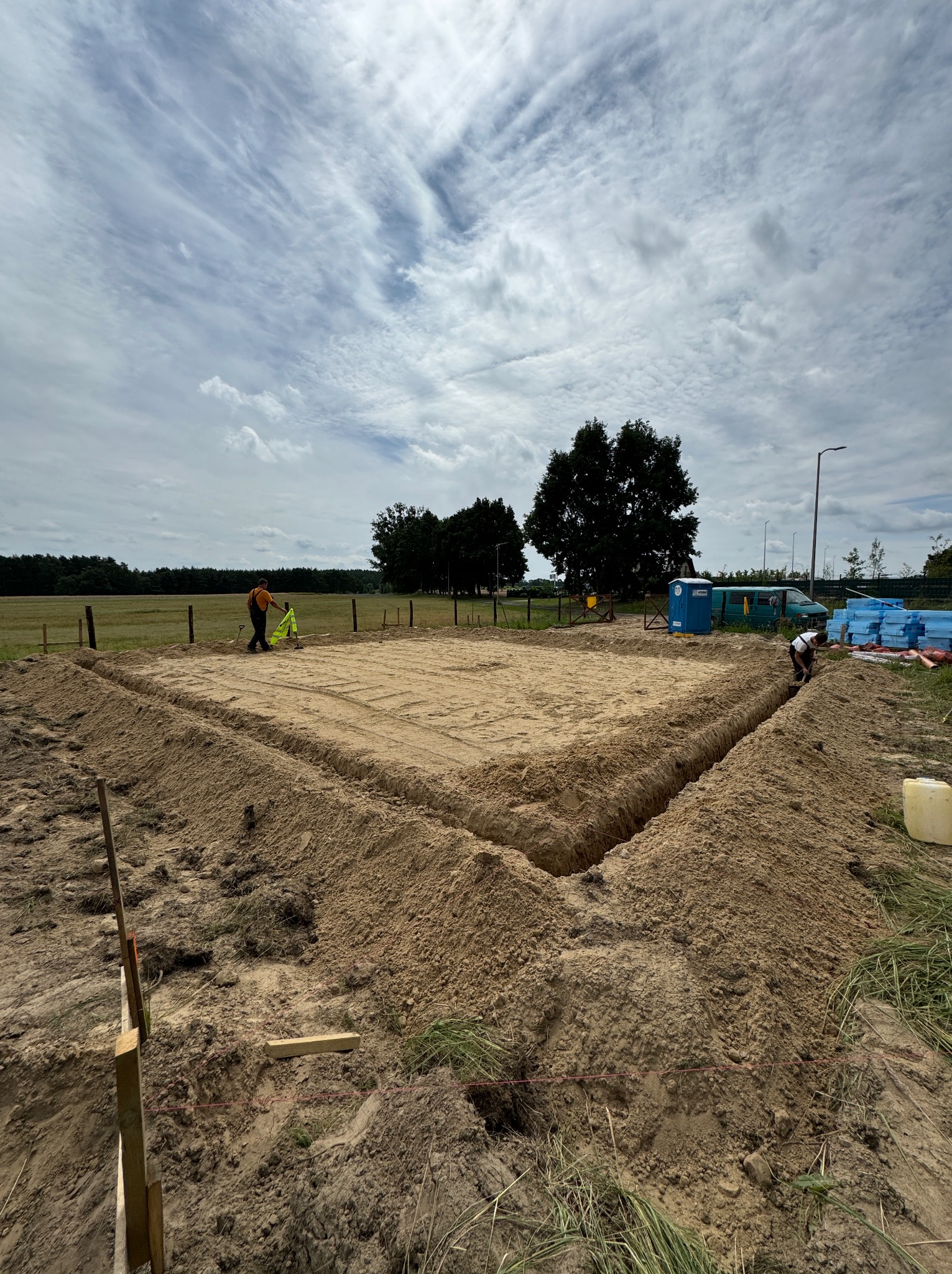Przygotowanie terenu pod fundamenty w Szczecinie: wykopane rowy, wyrównana powierzchnia wewnątrz, pracownicy w oddali, błękitny przenośny WC i zielony bus w tle, pochmurne niebo.
