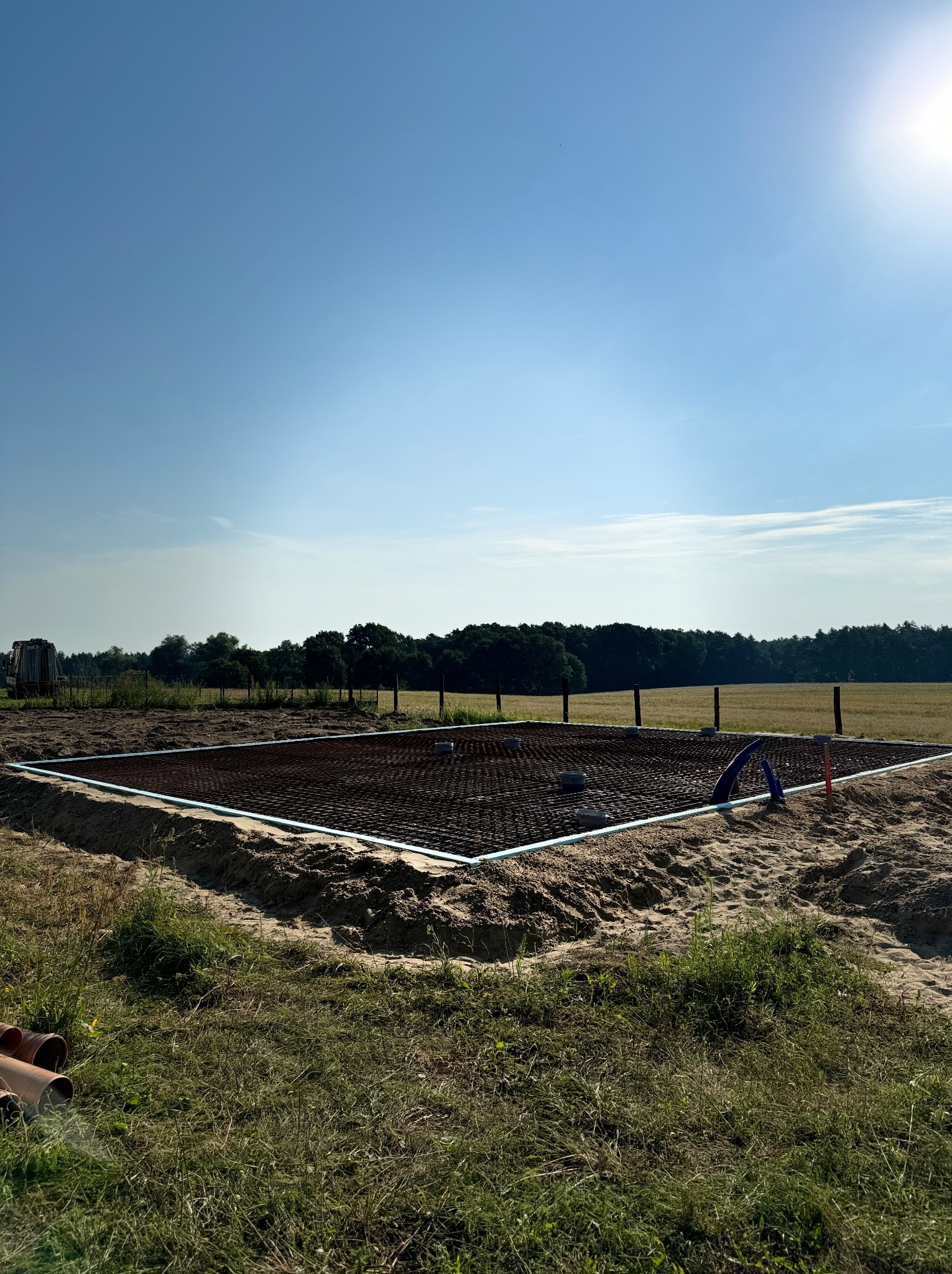 Przygotowany zbrojony szkielet pod wylewkę fundamentów, otoczony piaskiem i trawą, widok w słoneczny dzień.