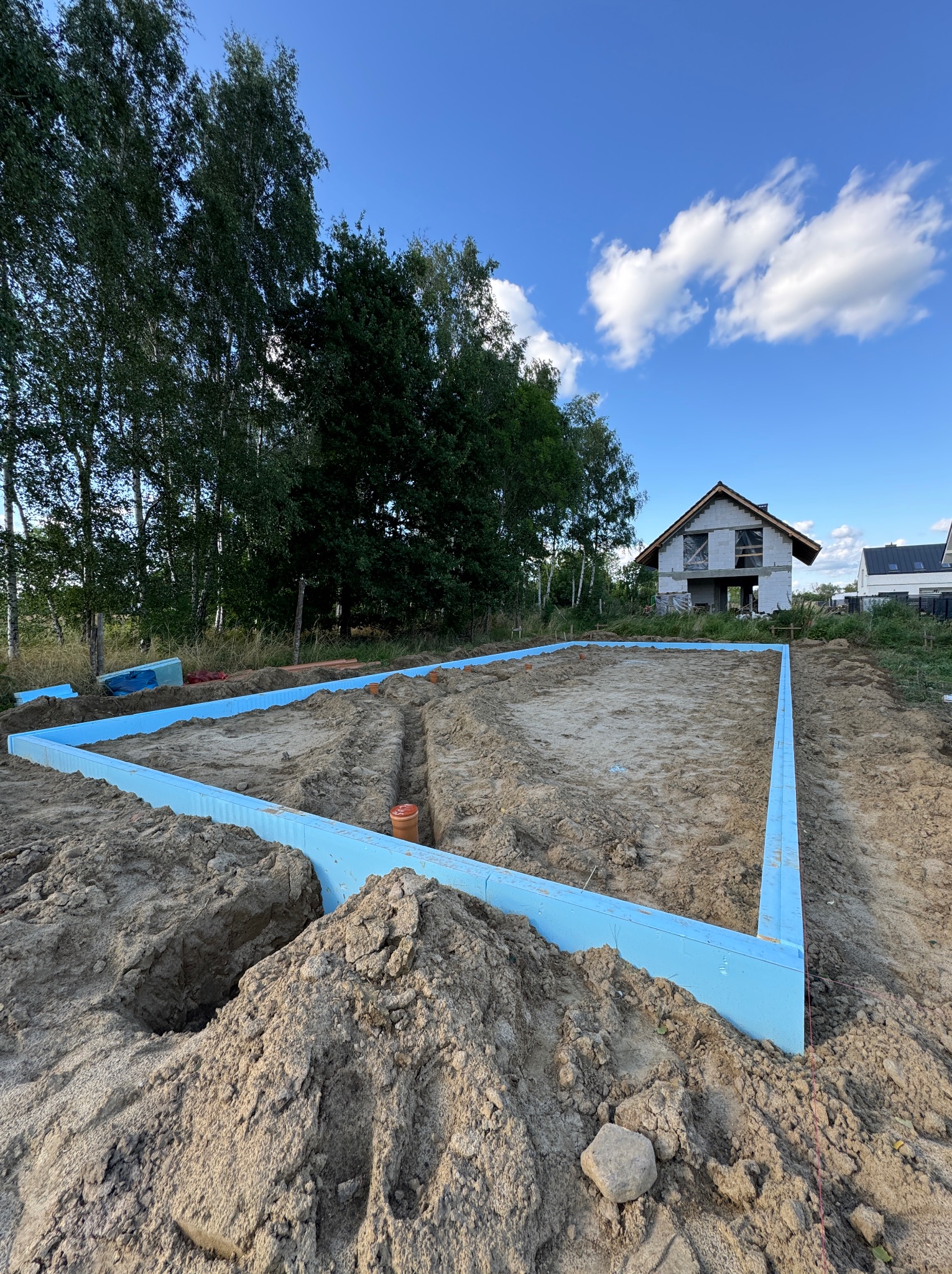 Przygotowany teren pod budowę domu jednorodzinnego z widocznym obrysem ławy fundamentowej wykonanym z niebieskiego styroduru, wykopane rowy na instalacje, w tle budynek w stanie surowym otwartym...