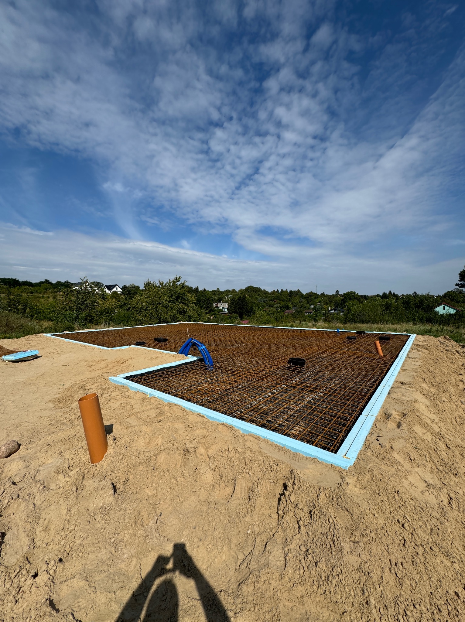 Przygotowany zbrojony szkielet fundamentów na piasku, obłożony niebieskim styropianem, z widocznymi rurami instalacyjnymi i shadow selfie na tle błękitnego nieba