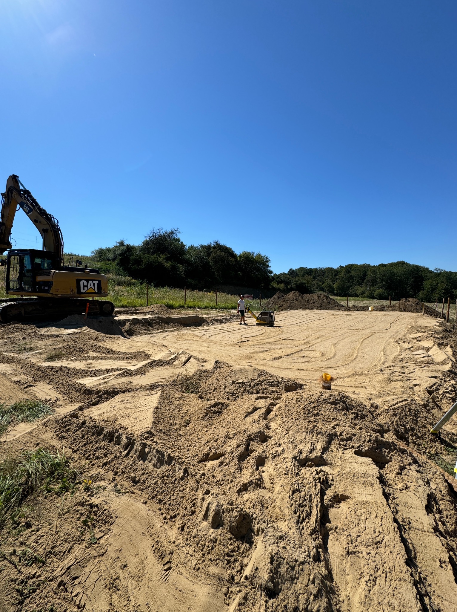 Przygotowanie terenu pod budowę: wykop z piaskiem, ubijarka wibracyjna i koparka CAT w tle pod błękitnym niebem.
