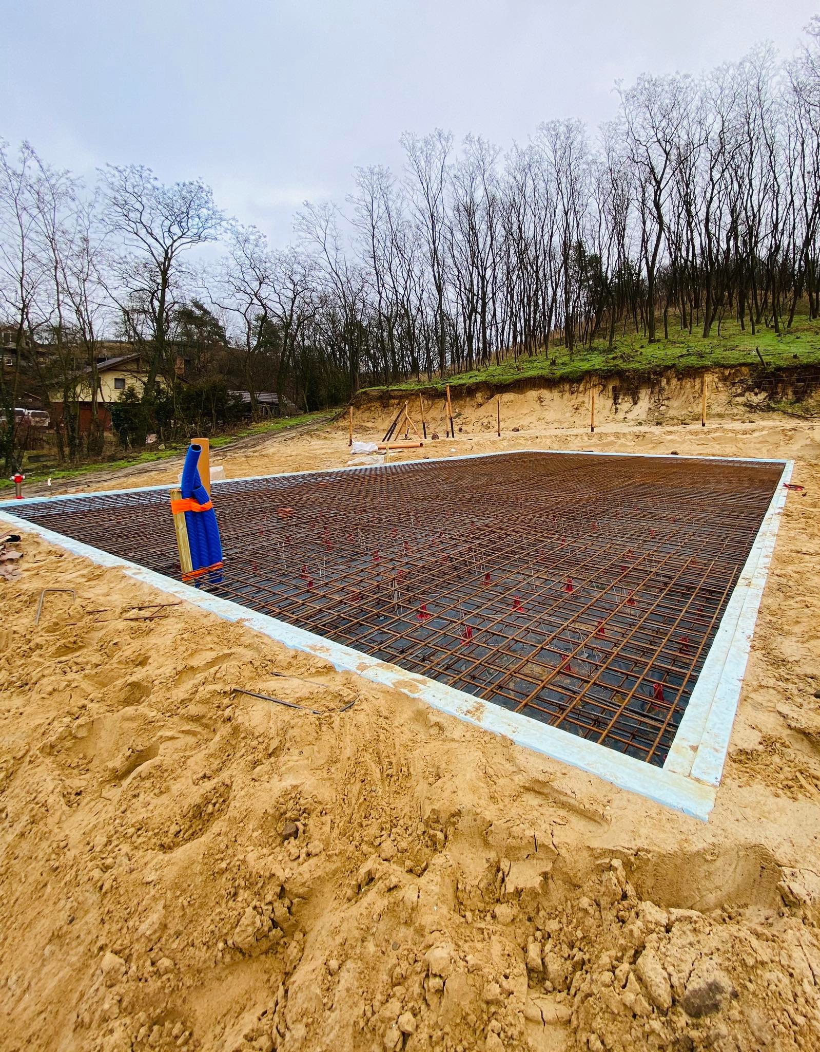 Przygotowany zbrojony wykop pod płytę fundamentową w Szczecinie, z widocznymi elementami szalunku i wystającymi prętami zbrojeniowymi, na tle piaszczystego terenu i drzew.