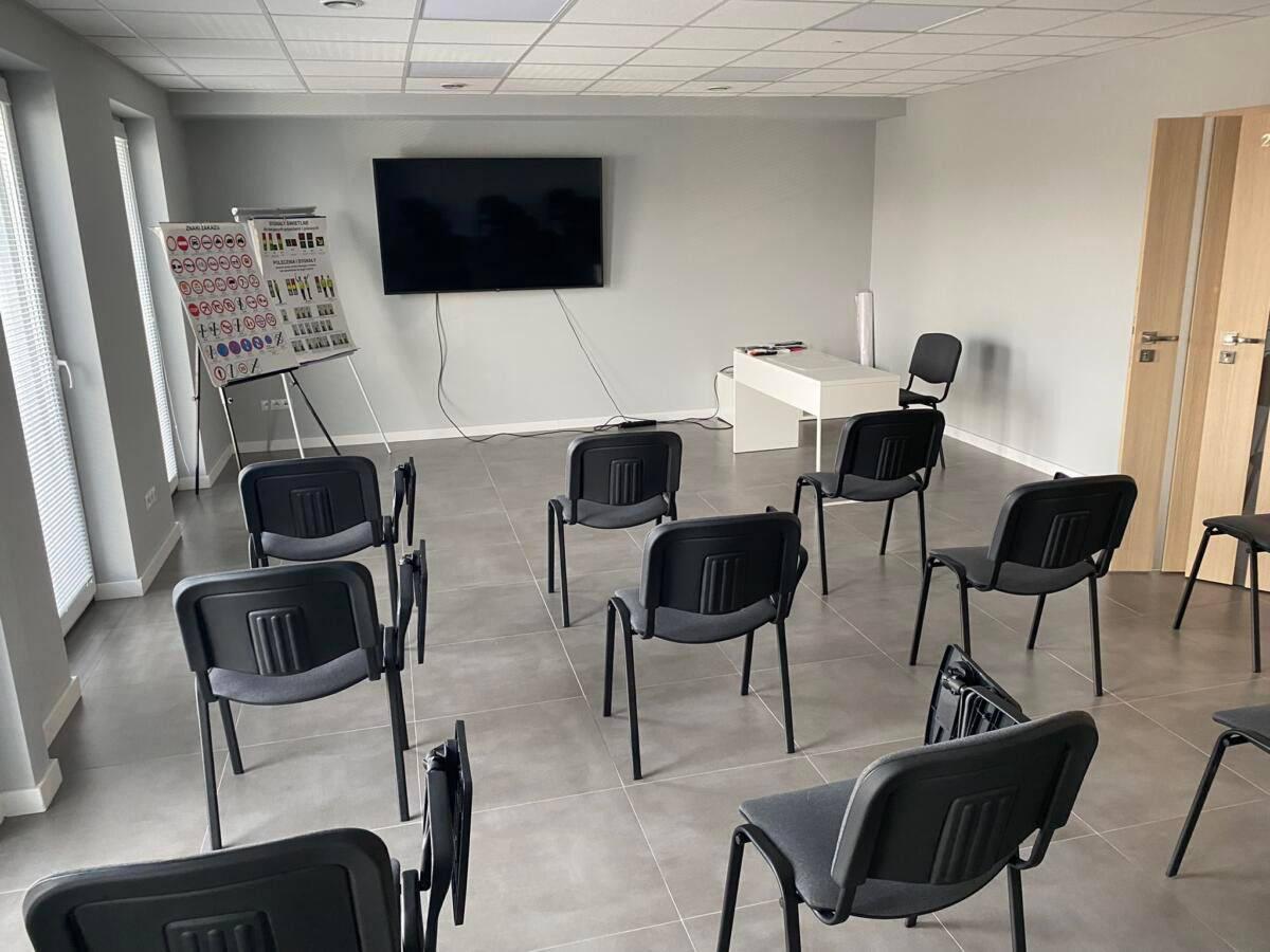 Sala wykładowa z krzesłami ustawionymi w rzędach, tablicą z zasadami ruchu drogowego na sztalugach, biurkiem i telewizorem na ścianie.