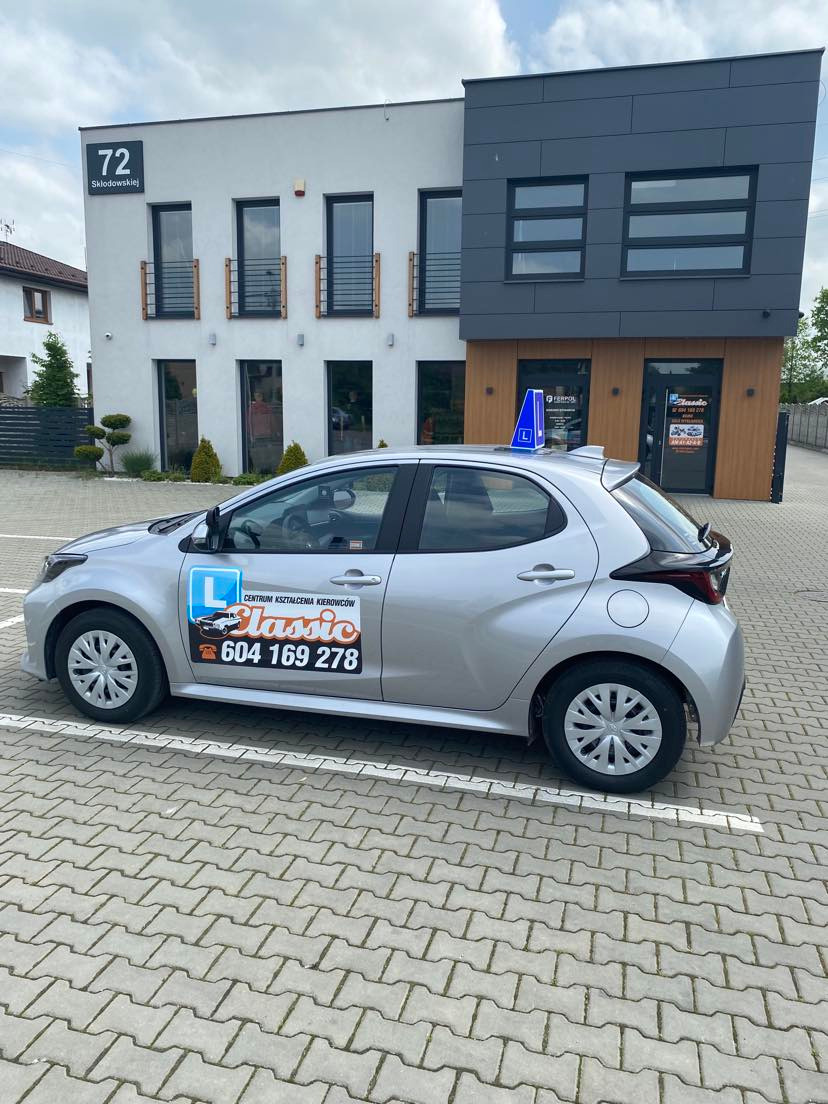 Srebrny samochód z oznaczeniem 'L' na dachu i naklejką z nazwą i numerem telefonu centrum kształcenia kierowców 'Classic' zaparkowany przed nowoczesnym budynkiem z numerem 72.