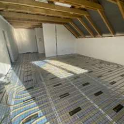 Żółte rury ogrzewania podłogowego ułożone w przestronnym pokoju na poddaszu, widoczne drewniane belki stropowe, przygotowane do wylewki.