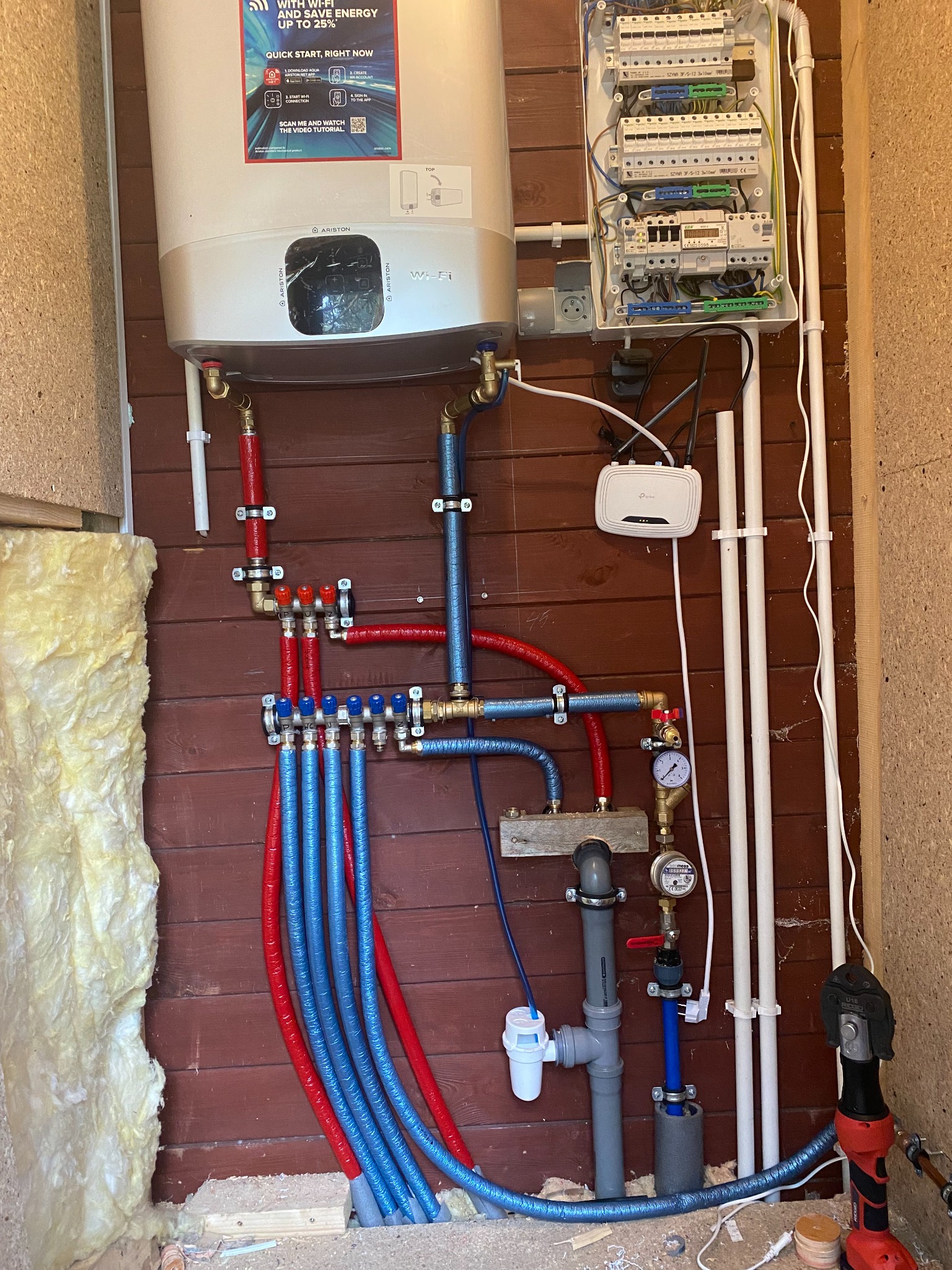 Węzeł cieplny w kotłowni z rozdzielaczem, rurami w izolacji czerwonej i niebieskiej, zaworami, manometrami, filtrem wody, piecem Ariston z WiFi, rozdzielnicą elektryczną i routerem TP-Link.