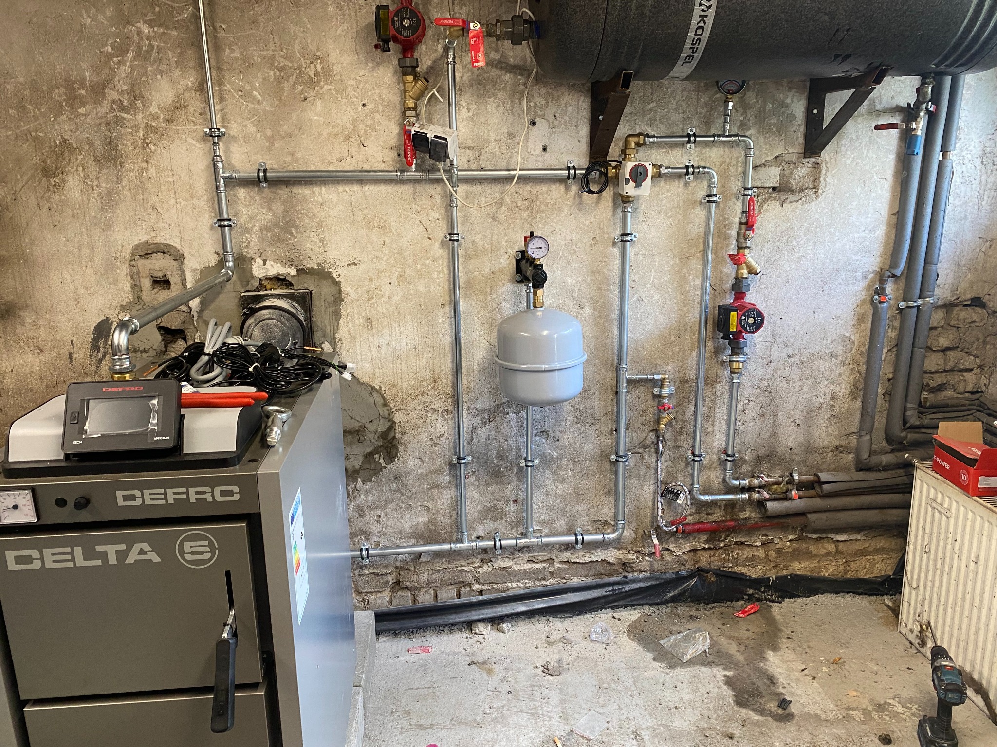 Nowa instalacja grzewcza z piecem Defro Delta 5, rurami stalowymi i zbiornikiem Kospel zamontowana na tle surowej, starej ściany z cegły i betonu.