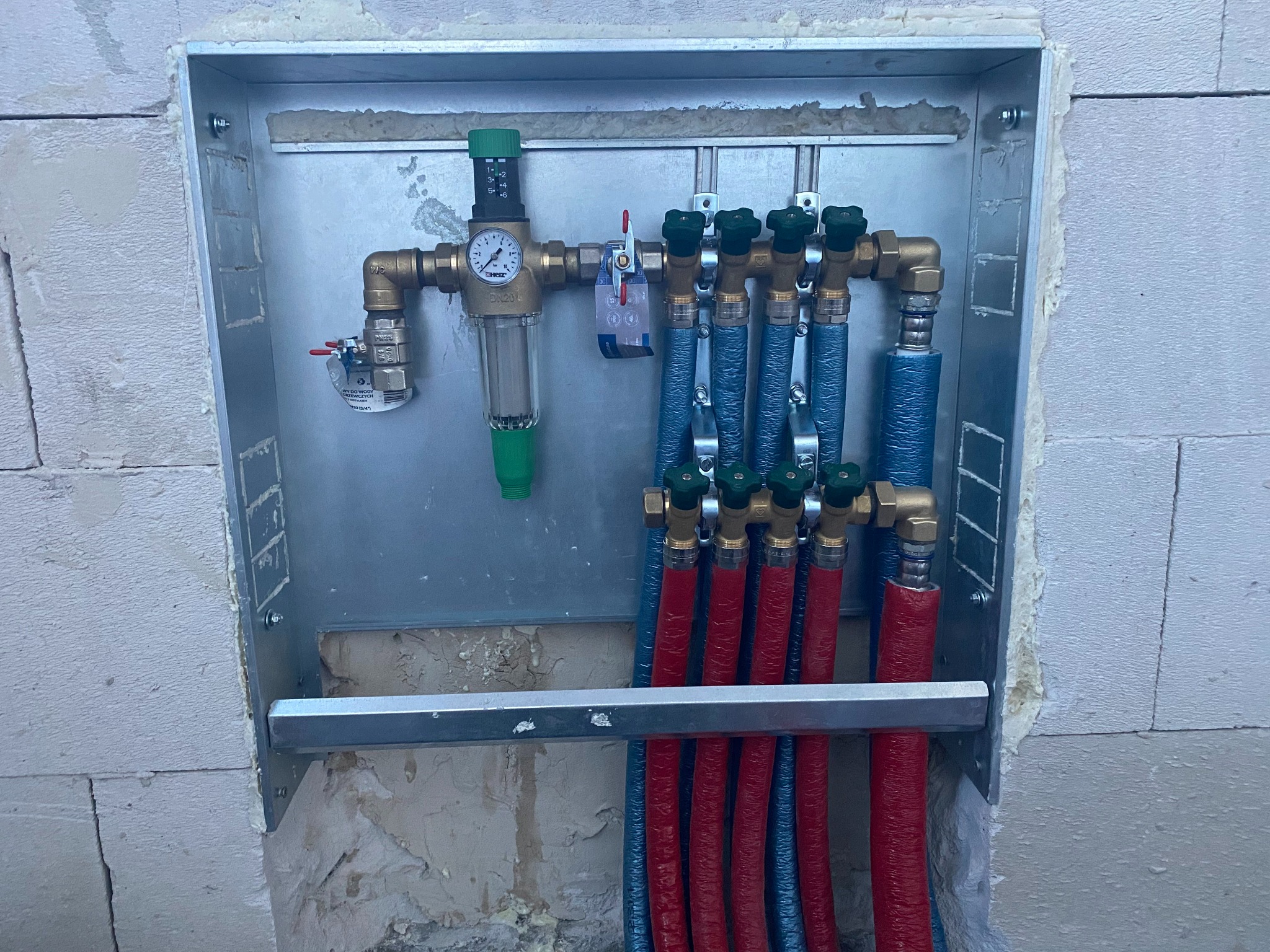 Rozdzielacz ogrzewania podłogowego w metalowej szafce, widoczne rury w izolacji niebieskiej i czerwonej, manometr, filtr i zawory, zamontowany w surowej ścianie z betonu komórkowego.