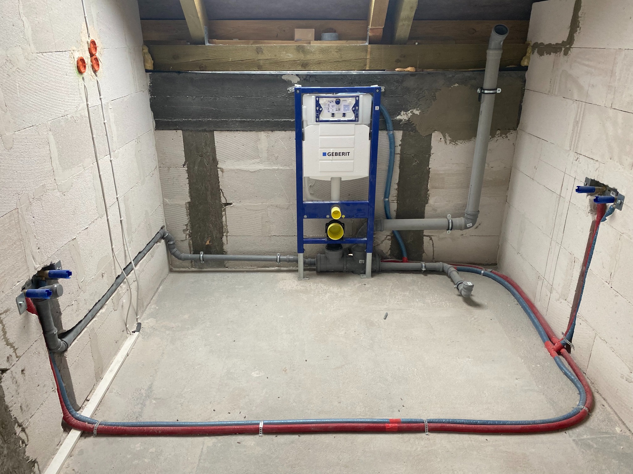 Stelaż podtynkowy Geberit zamontowany w surowym pomieszczeniu z widocznymi instalacjami wodno-kanalizacyjnymi, rury prowadzone po podłodze, ściany z białego bloczka.