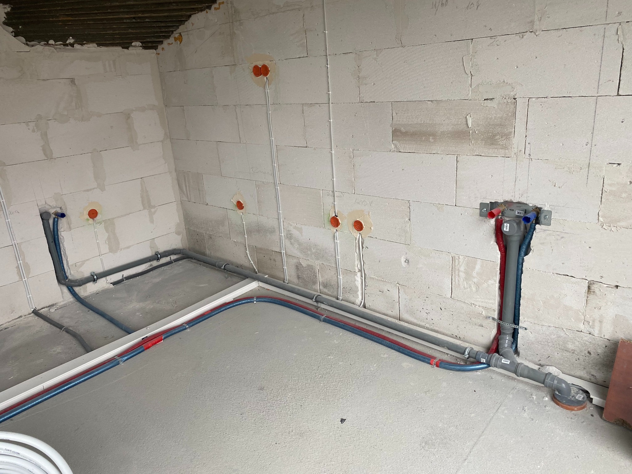 Surowy stan instalacji wodno-kanalizacyjnej w trakcie budowy, widoczne rury PEX w kolorze czerwonym i niebieskim prowadzone po posadzce, szare rury kanalizacyjne w ścianie z betonu komórkowego...