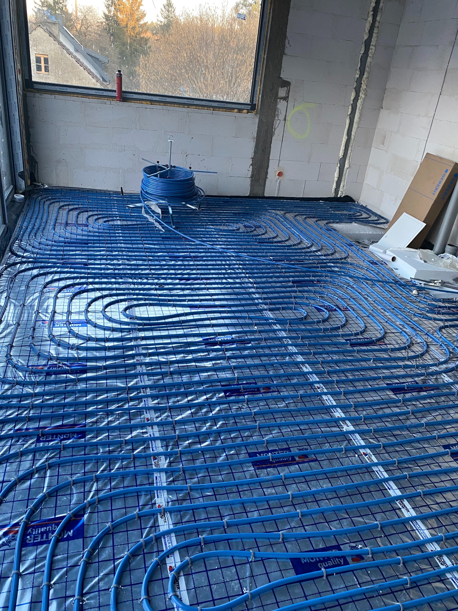 Niebieskie rury ogrzewania podłogowego ułożone na folii z nadrukiem, zamocowane do siatki w surowym stanie, widok na okno z widokiem na drzewa i budynek.