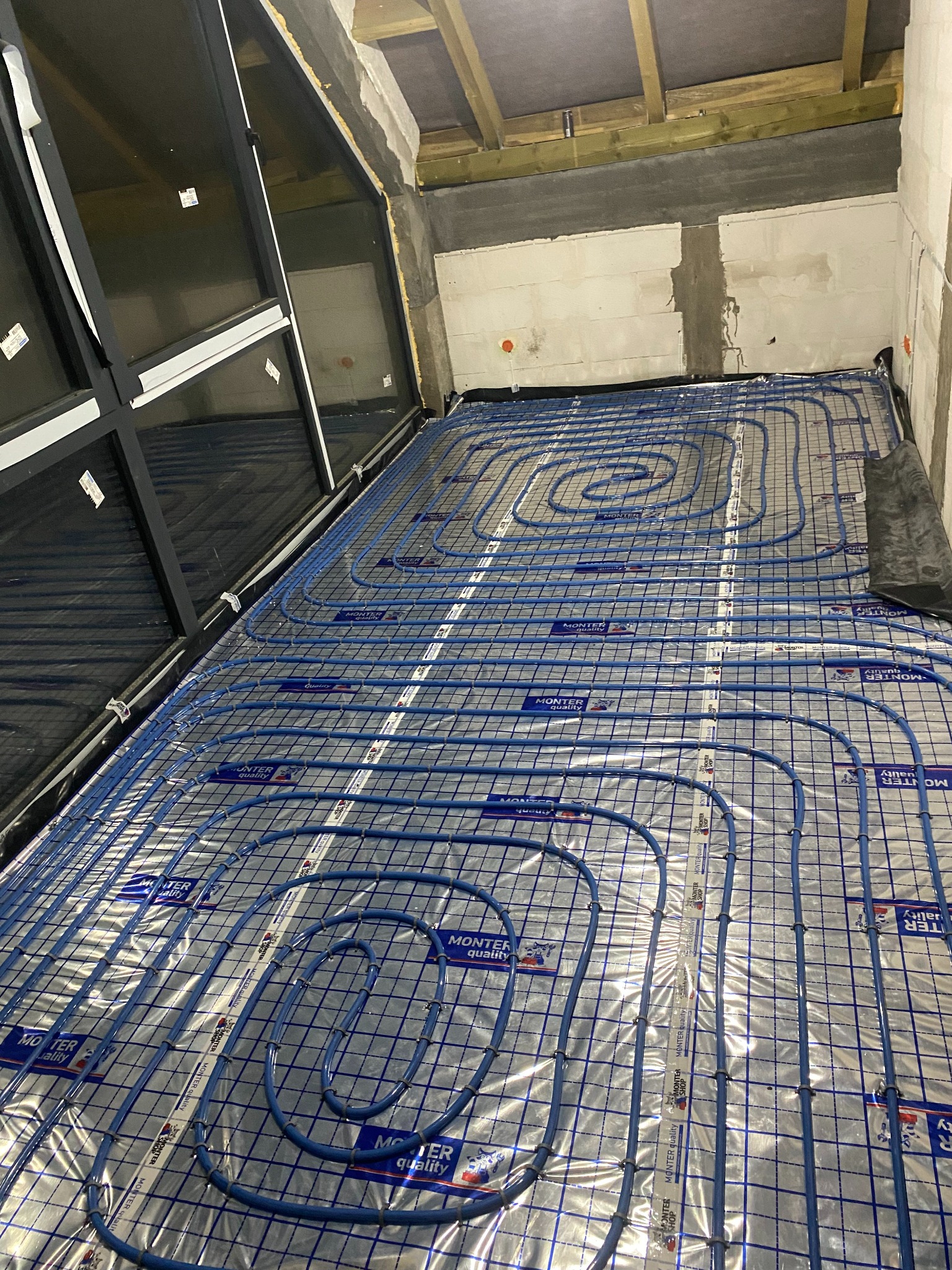 Niebieskie rury ogrzewania podłogowego ułożone na srebrnej folii z nadrukiem w kratkę, widok z góry w surowym wnętrzu z drewnianą konstrukcją dachu i ścianami z betonu komórkowego.