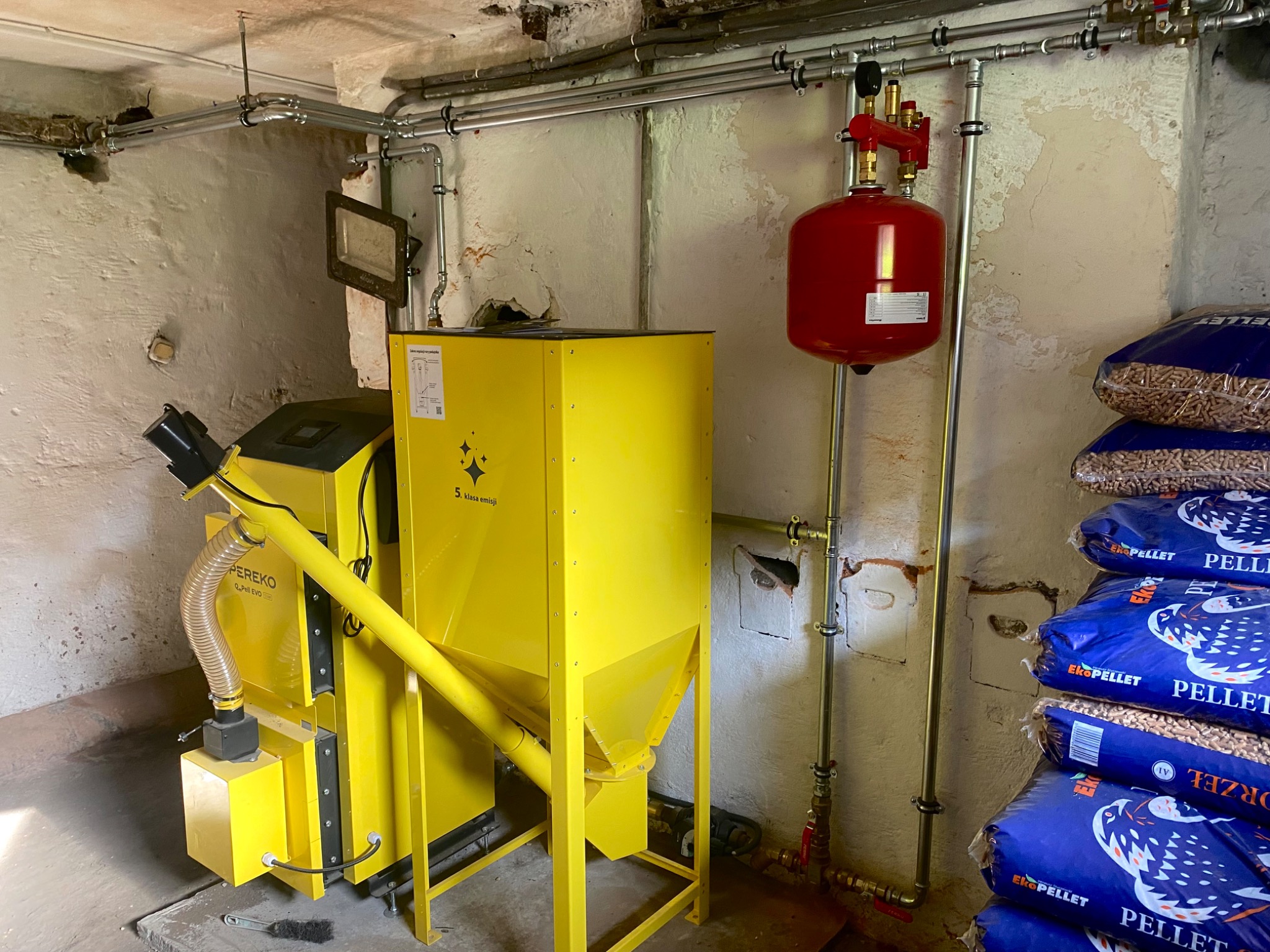 Żółty piec na pellet marki PEREKO w kotłowni, z czerwoną instalacją i zapasem pelletu w workach. Widoczne rury instalacji grzewczej.