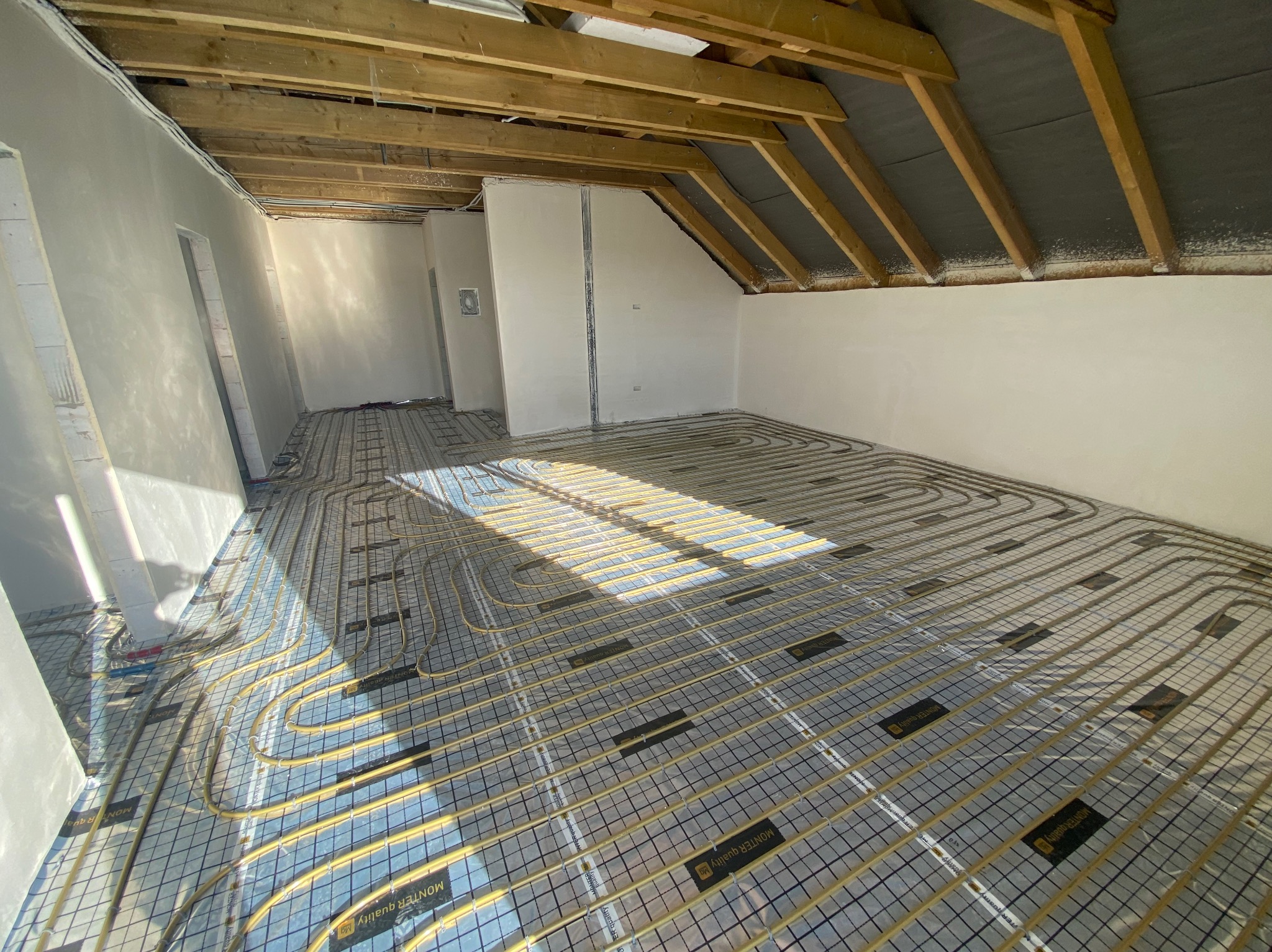 Żółte rury ogrzewania podłogowego ułożone w przestronnym pokoju na poddaszu, widoczne drewniane belki stropowe, przygotowane do wylewki.