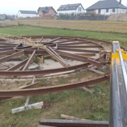 Konstrukcja z metalowych profili w kształcie okręgu z promieniami, w trakcie montażu na trawiastym terenie, widoczny dźwig i spawarka, w tle zabudowa jednorodzinna.