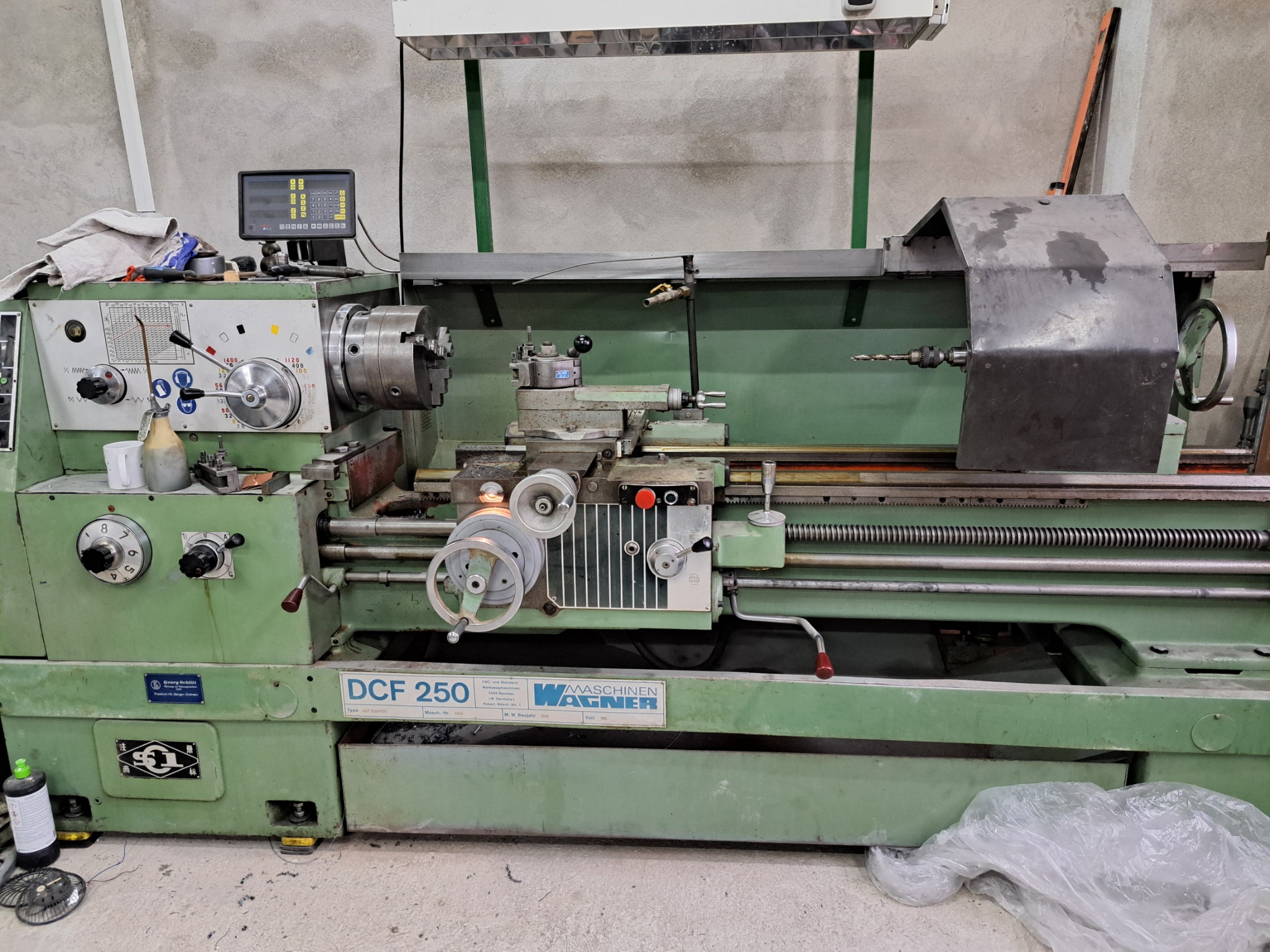 Zielona tokarka uniwersalna DCF 250 z widocznym wyświetlaczem, uchwytem tokarskim i zestawem narzędzi, oświetlona lampą warsztatową, na betonowej posadzce.