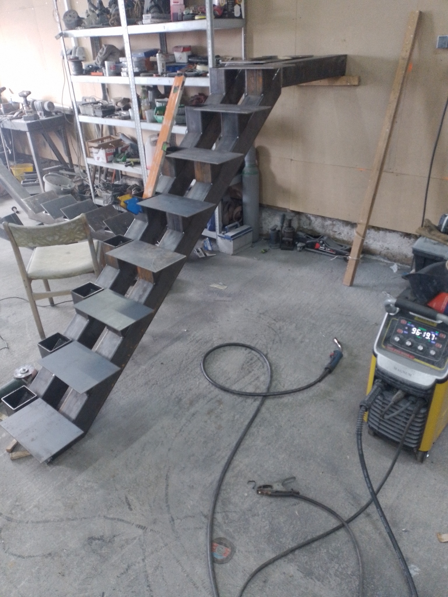 Konstrukcja schodów metalowych w trakcie spawania, widoczna spawarka i narzędzia w warsztacie.