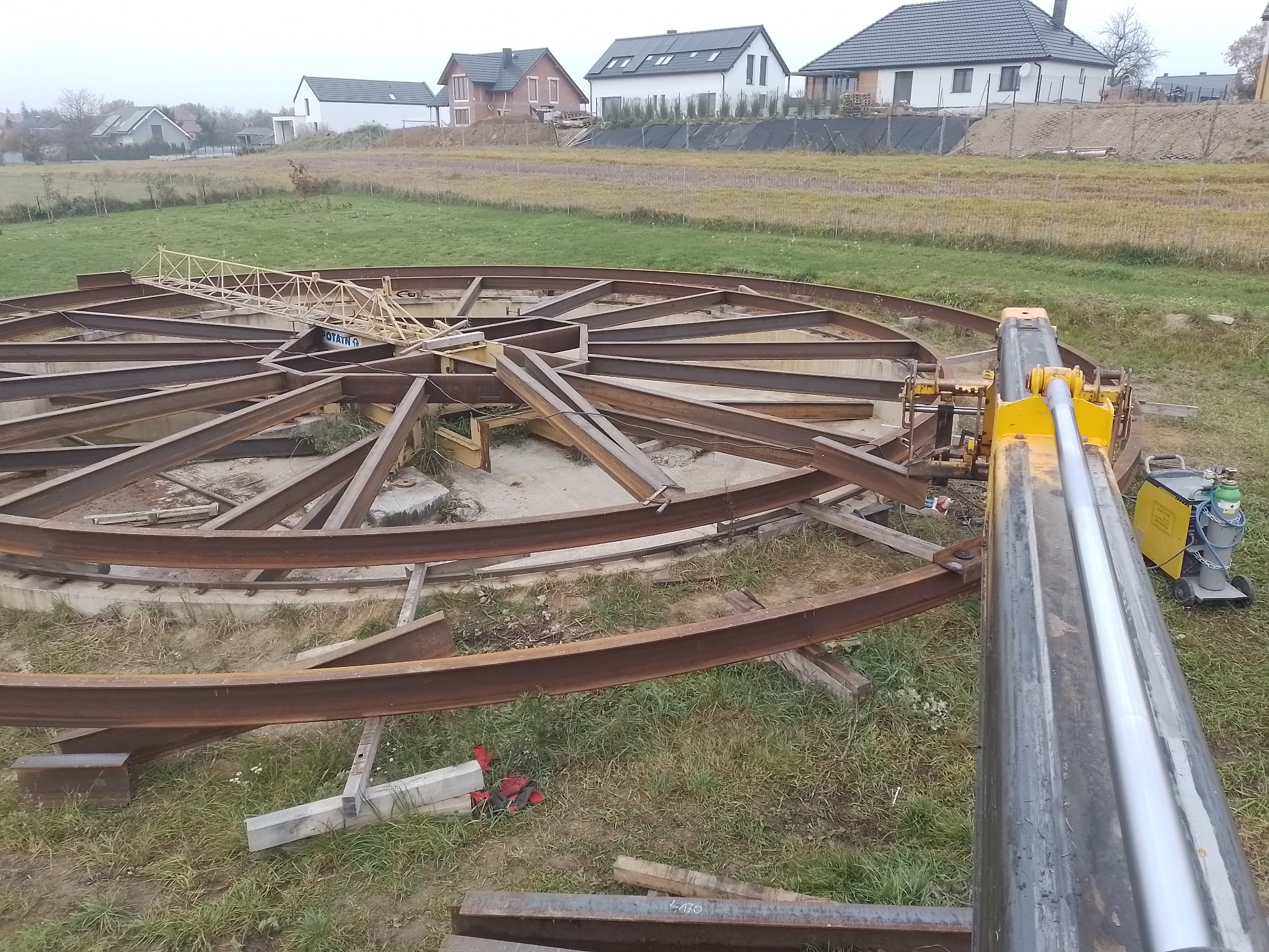 Konstrukcja z metalowych profili w kształcie okręgu z promieniami, w trakcie montażu na trawiastym terenie, widoczny dźwig i spawarka, w tle zabudowa jednorodzinna.