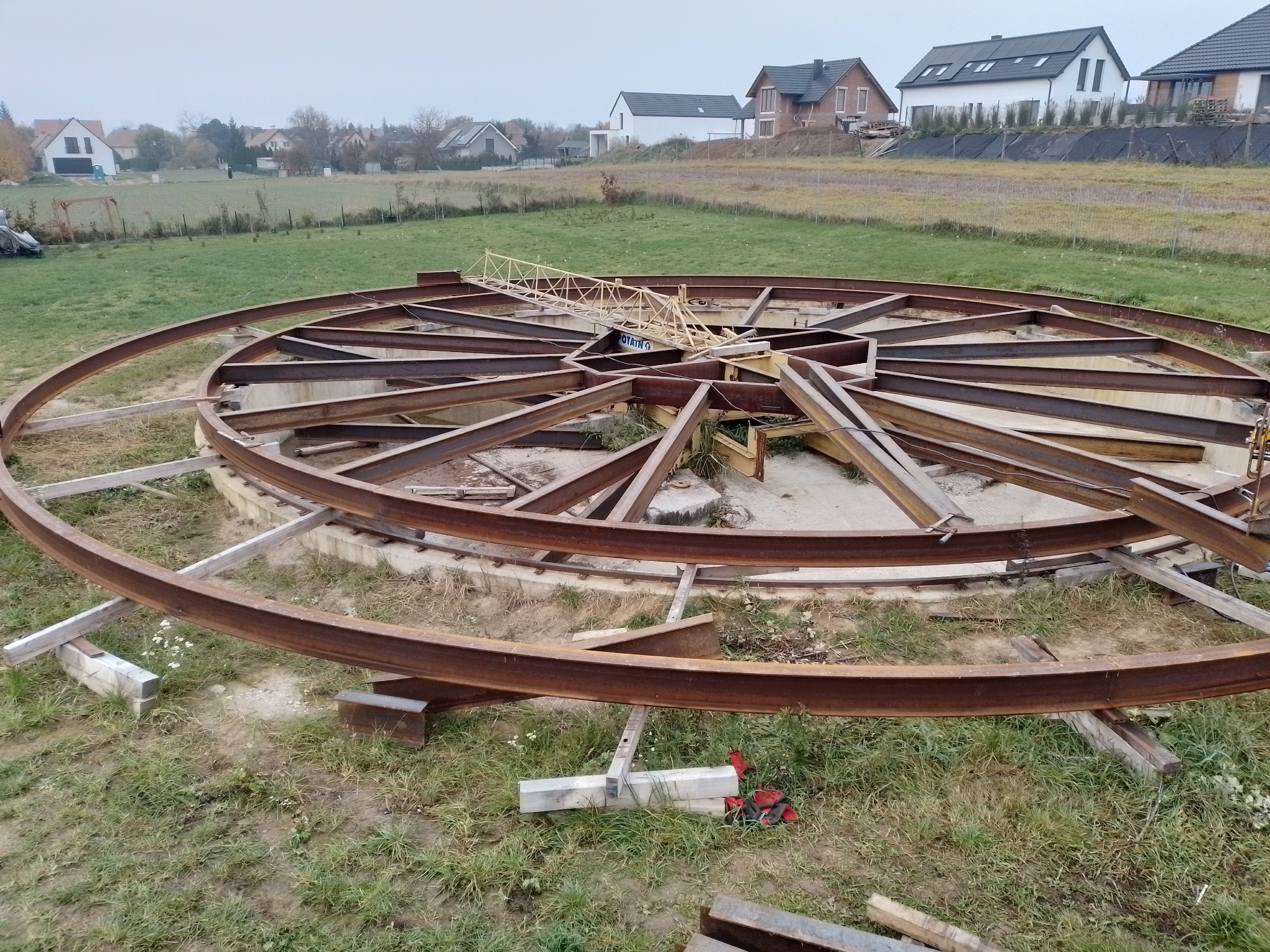 Konstrukcja z metalowych belek w kształcie okręgu, prawdopodobnie pod fundament, z fragmentem żurawia wieżowego wewnątrz, na tle domów jednorodzinnych i pola.