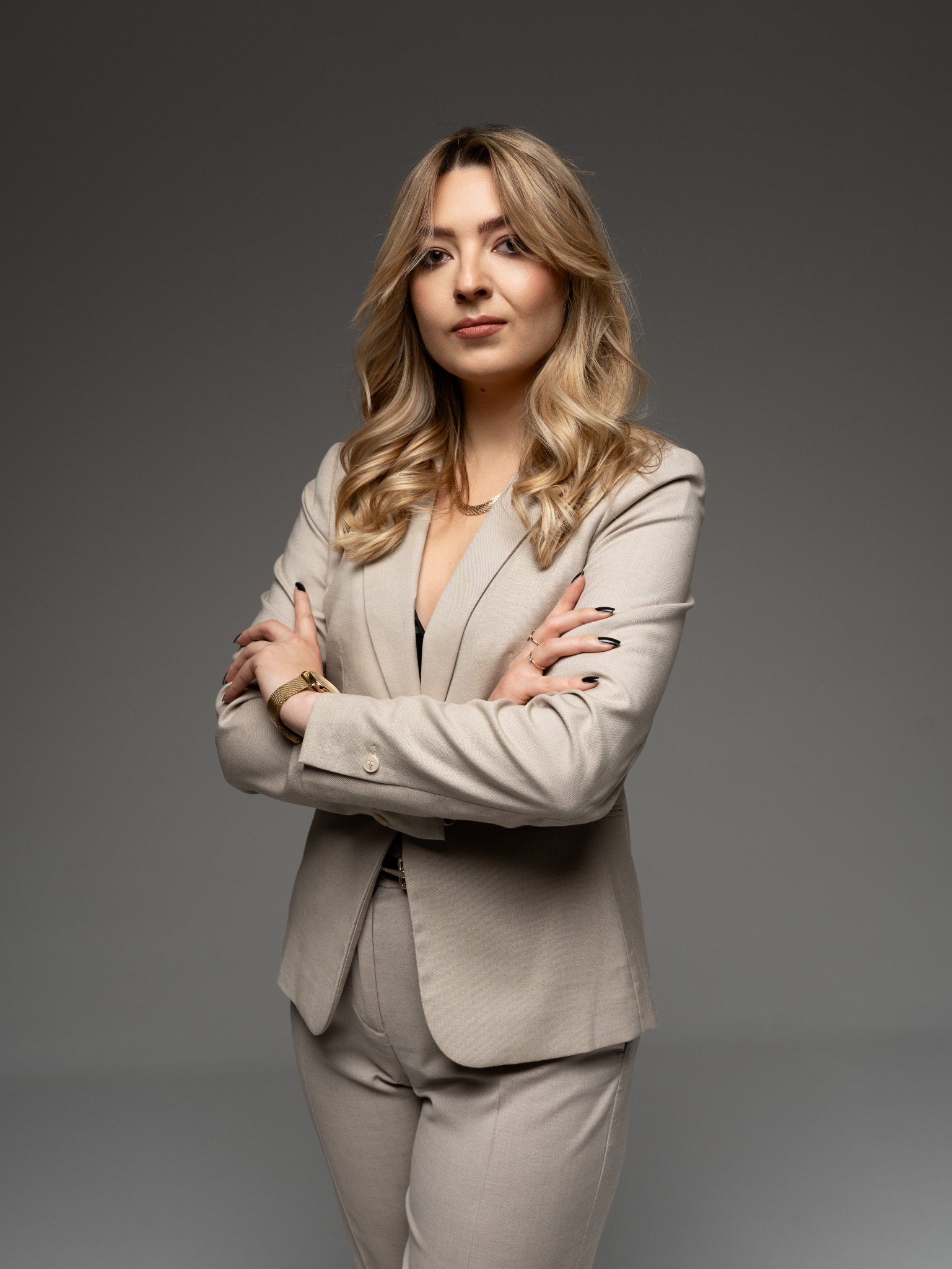Portret kobiety w beżowym garniturze, z założonymi rękami i złotym zegarkiem na tle szarego studia.