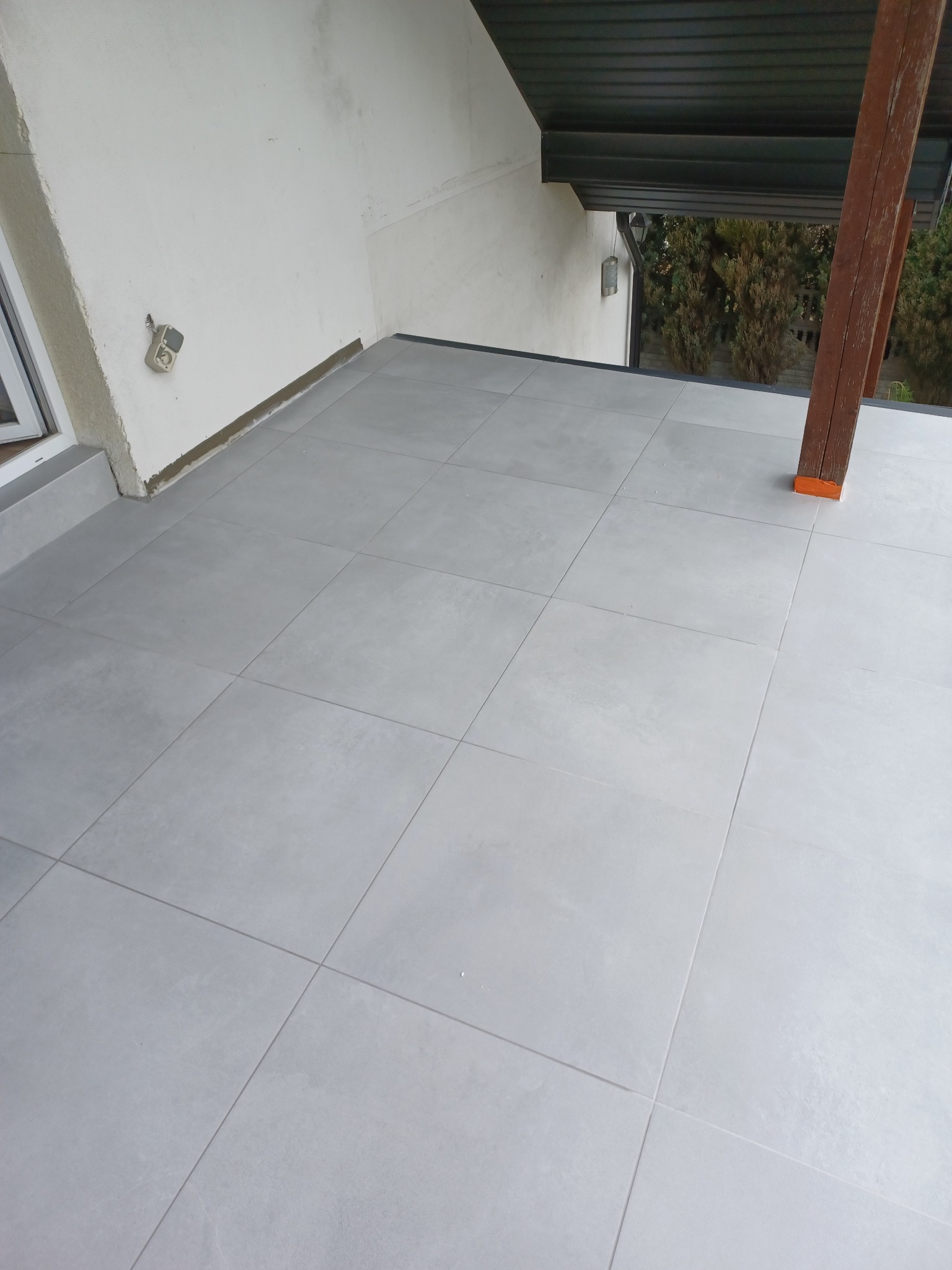 Jasnoszara, kwadratowa terakota na zadaszonym tarasie, widoczny fragment drewnianej kolumny i zewnętrzna ściana budynku z gniazdkiem elektrycznym.