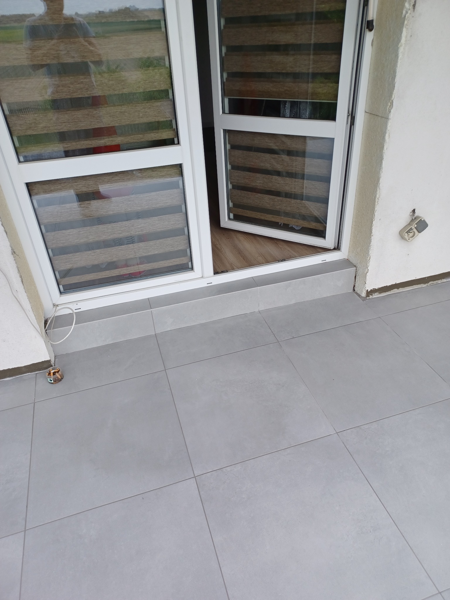 Szare płytki podłogowe na balkonie, z widocznym oknem balkonowym z żaluzjami poziomymi i niedokończonym gniazdkiem elektrycznym na ścianie.