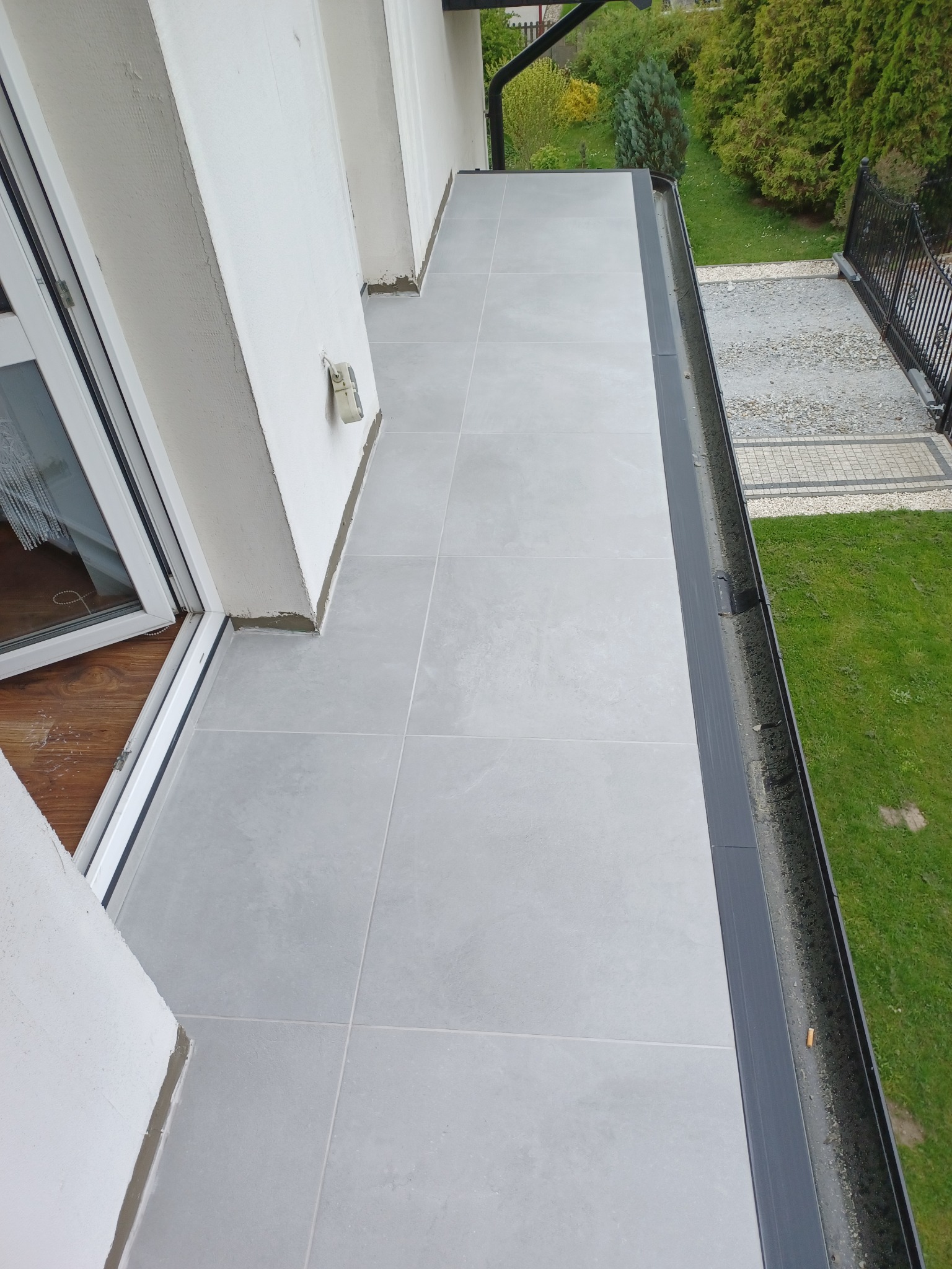 Szare płytki gresowe na balkonie, widok z góry, z widocznym fragmentem drzwi balkonowych i ogrodu w tle.