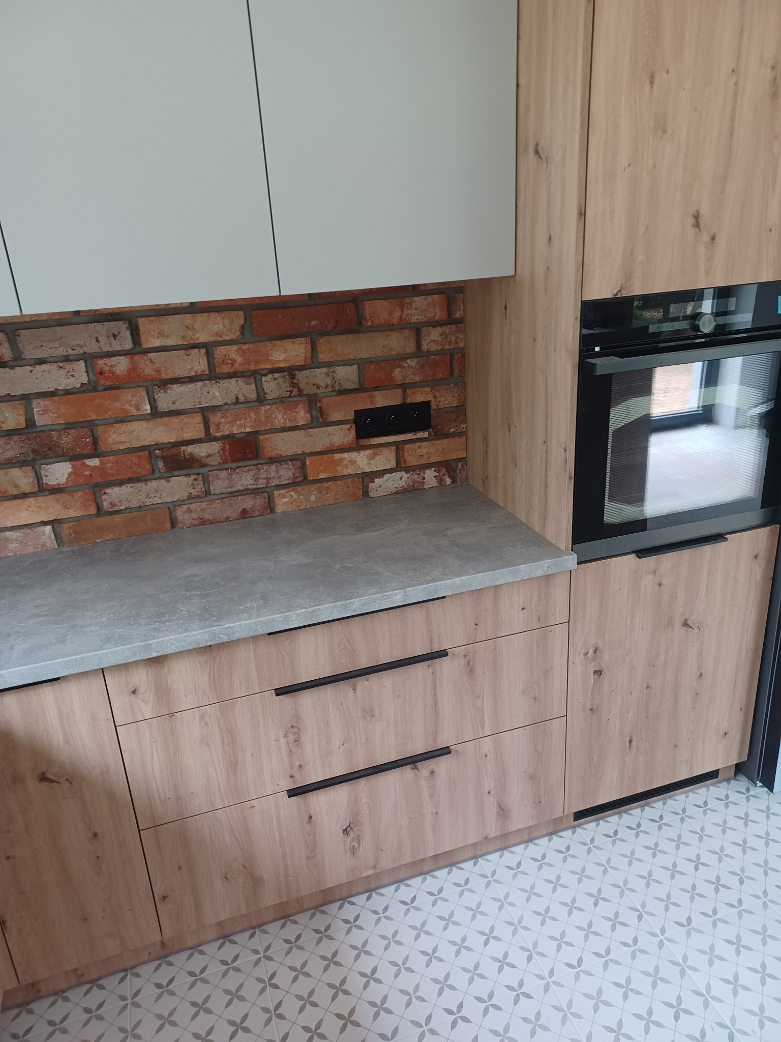 Fragment zabudowy kuchennej: drewnopodobne fronty szafek, szary blat, ceglana ściana nad blatem, piekarnik w zabudowie, wzorzysta podłoga.