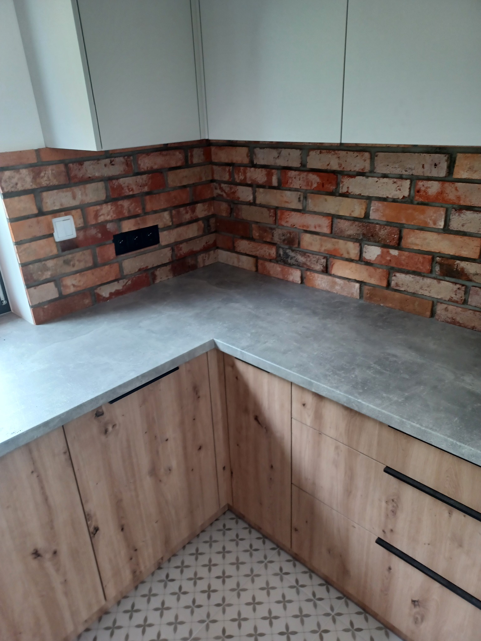 Kuchnia z ceglaną ścianą nad blatem, szary blat i drewniane szafki z czarnymi uchwytami, wzorzysta podłoga.