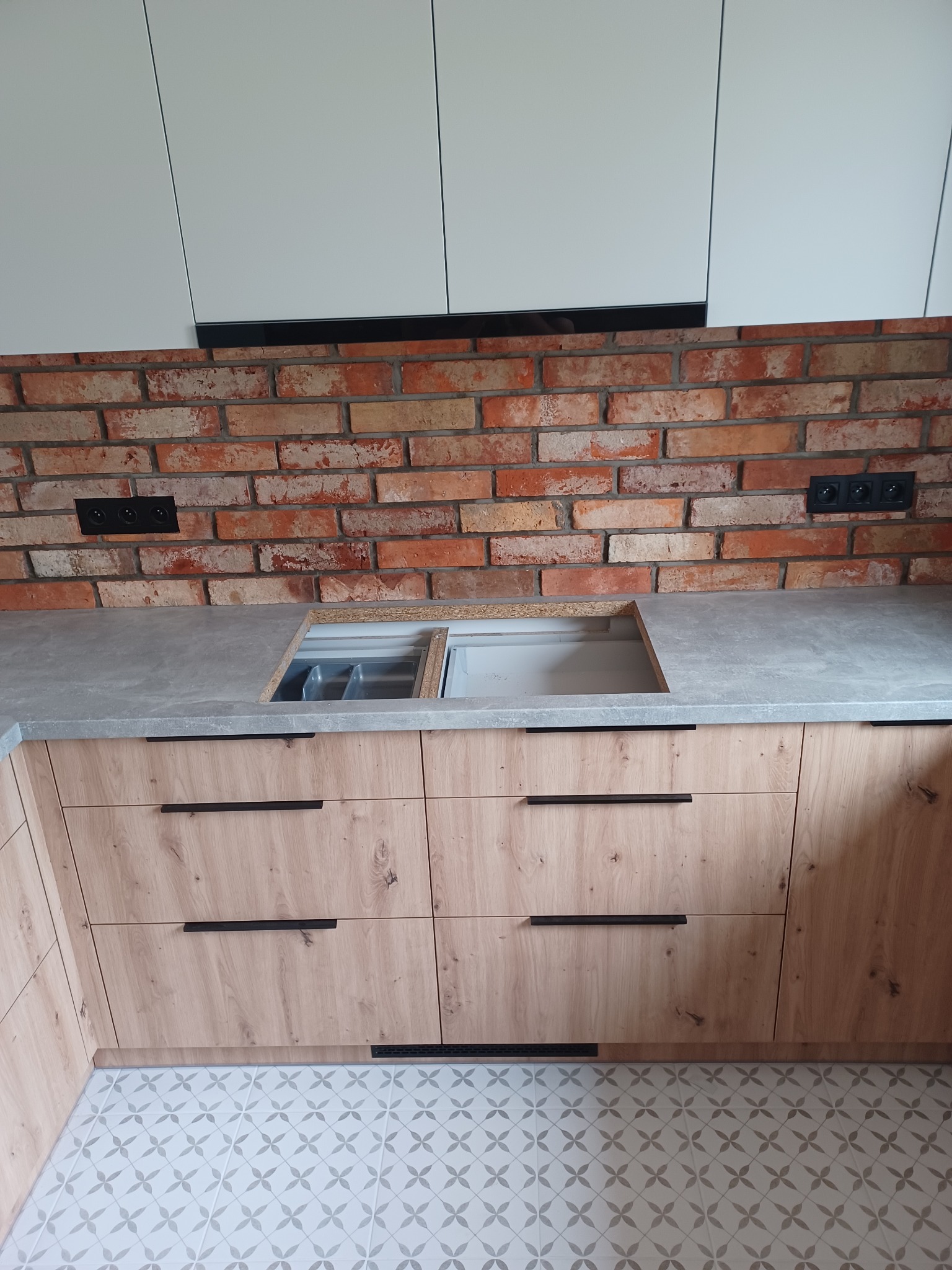 Fragment kuchni w trakcie montażu: drewnopodobne szafki z czarnymi uchwytami, szary blat z wyciętym otworem na zlew, ceglana ściana z gniazdkami, białe szafki górne i wzorzysta podłoga.