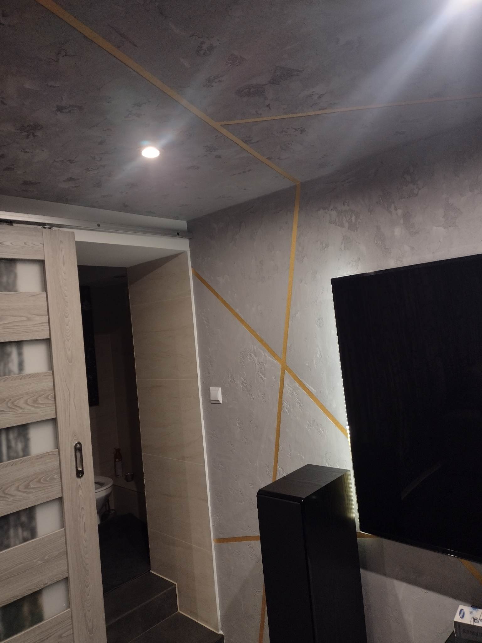 Szara ściana i sufit z dekoracyjnymi złotymi paskami, widoczny fragment przesuwanych drzwi do łazienki i fragment czarnego telewizora oraz głośnika.