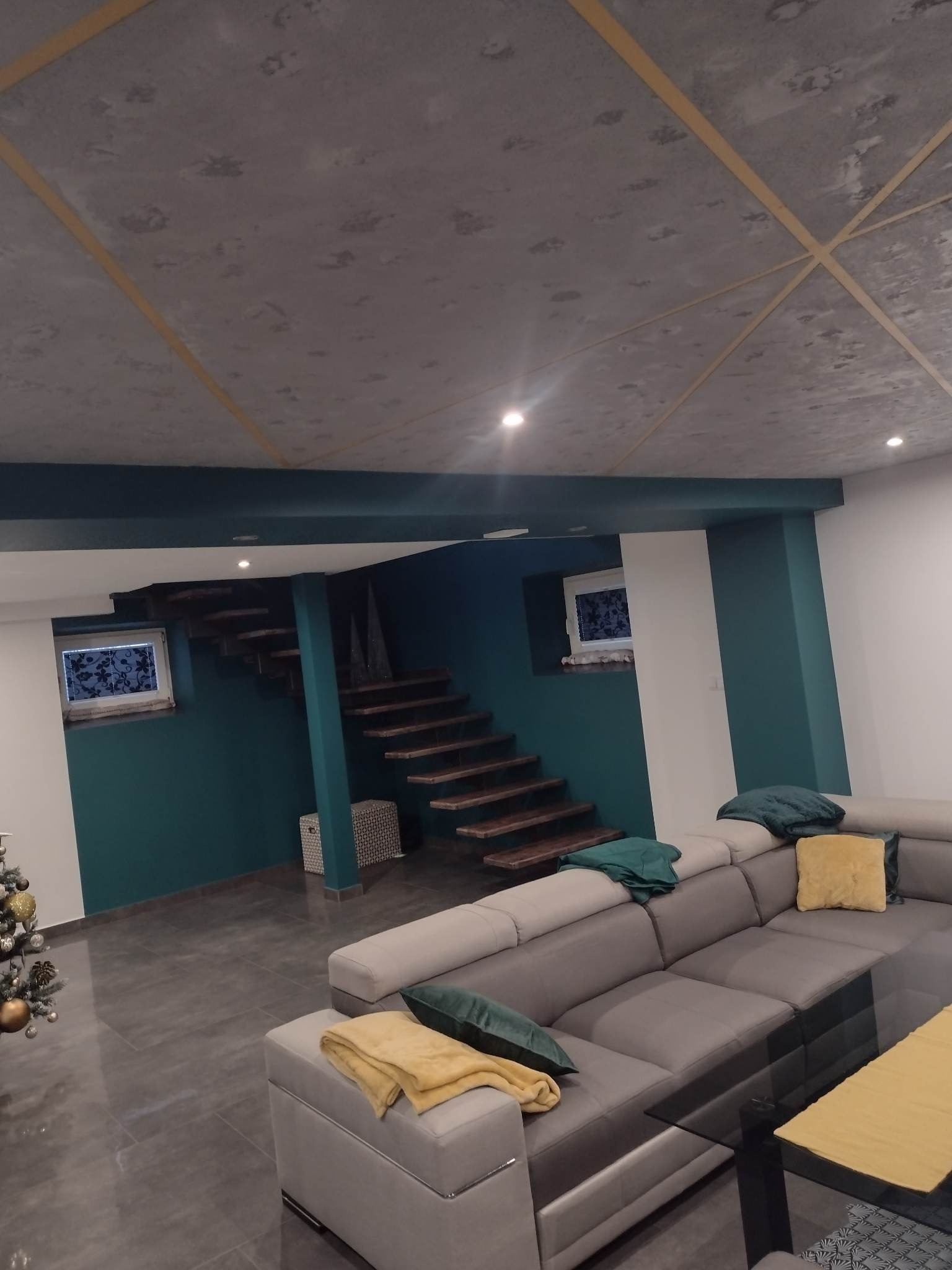 Nowoczesny salon z drewnianymi schodami, szarą sofą narożną z poduszkami i kocem w odcieniach żółci oraz sufitem z widoczną strukturą betonu i dekoracyjnymi listwami.