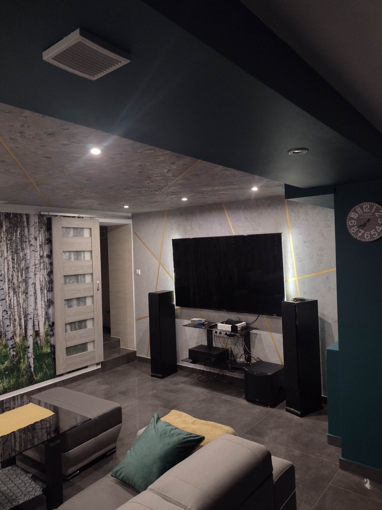 Nowoczesny salon z szarą tapetą w geometryczne wzory, ciemnozielonym sufitem, przesuwnymi drzwiami z motywem brzozowego lasu oraz czarnym sprzętem audio-wideo.