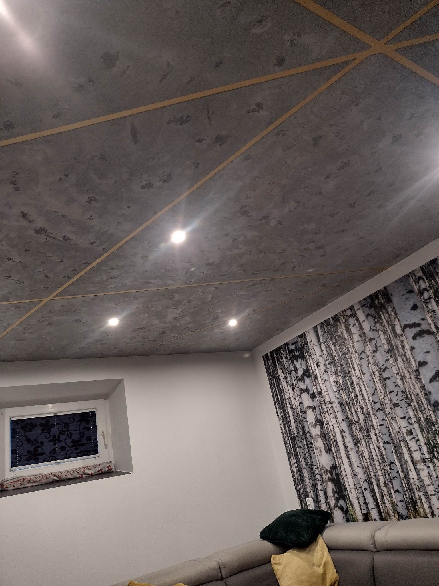Betonowy sufit z geometrycznym podziałem listwami, oświetleniem punktowym i tapetą z motywem brzozowego lasu na ścianie