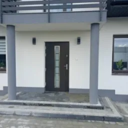 Wejście do domu z nową kostką brukową, szara elewacja, stolarka okienna w kolorze brązowym, widoczna czerwona łopata oparta o ścianę.