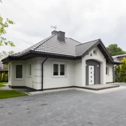 Elegancki, parterowy dom z białą elewacją, szarą kostką brukową na podjeździe i zadbanym trawnikiem, widok z zewnątrz w pochmurny dzień.