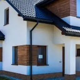 Nowoczesny dom jednorodzinny z ocieploną i pomalowaną elewacją w kolorze białym, z elementami drewnianymi w kolorze brązowym, czarną dachówką i rynnami, widoczne okna z roletami zewnętrznymi.