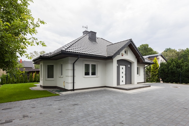 Elegancki, parterowy dom z białą elewacją, szarą kostką brukową na podjeździe i zadbanym trawnikiem, widok z zewnątrz w pochmurny dzień.