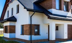 Nowoczesny dom jednorodzinny z ocieploną i pomalowaną elewacją w kolorze białym, z elementami drewnianymi w kolorze brązowym, czarną dachówką i rynnami, widoczne okna z roletami zewnętrznymi.