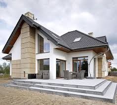 Nowoczesny dom jednorodzinny w Krosno z białą elewacją, fragmentami wykończonymi piaskowcem, dużymi oknami i tarasem z szarych płyt. Widoczne meble ogrodowe na tarasie.