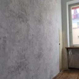 Ściana po nałożeniu dekoracyjnej szarej tapety o nieregularnym wzorze, widoczny parkiet na podłodze i fragment okna.