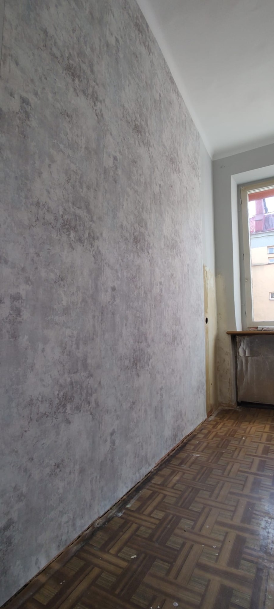 Ściana po nałożeniu dekoracyjnej szarej tapety o nieregularnym wzorze, widoczny parkiet na podłodze i fragment okna.