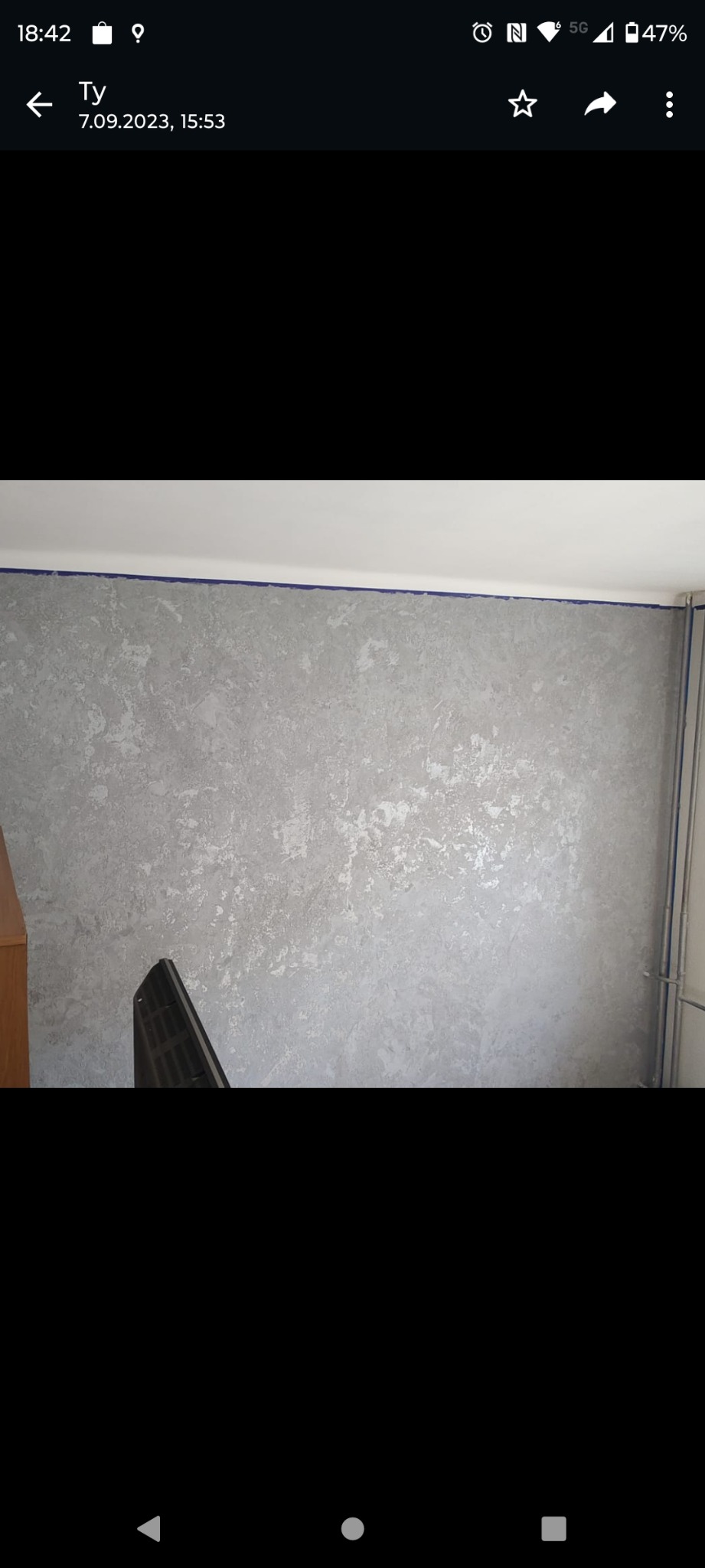 Ściana w odcieniach szarości z efektem dekoracyjnym, zabezpieczona niebieską taśmą malarską przy suficie, widoczne rury grzewcze.